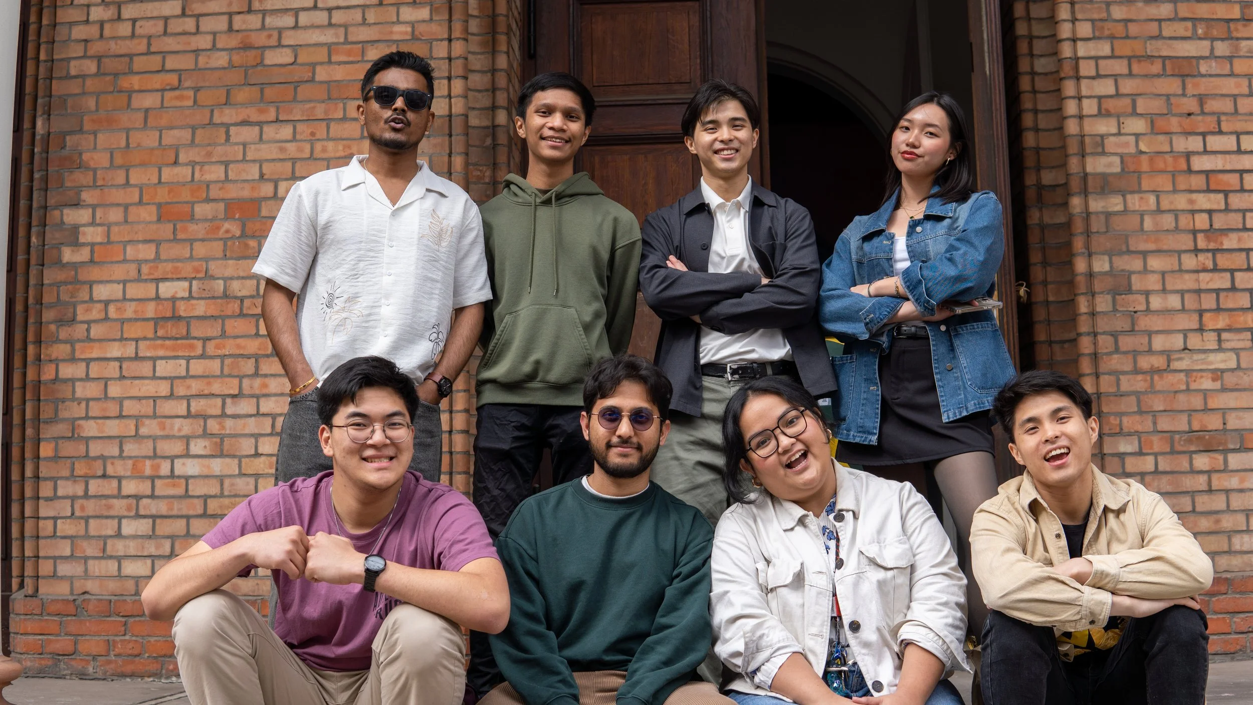Group of nine young adults standing and sitting outside in front of a brick building, smiling and posing for a photo.