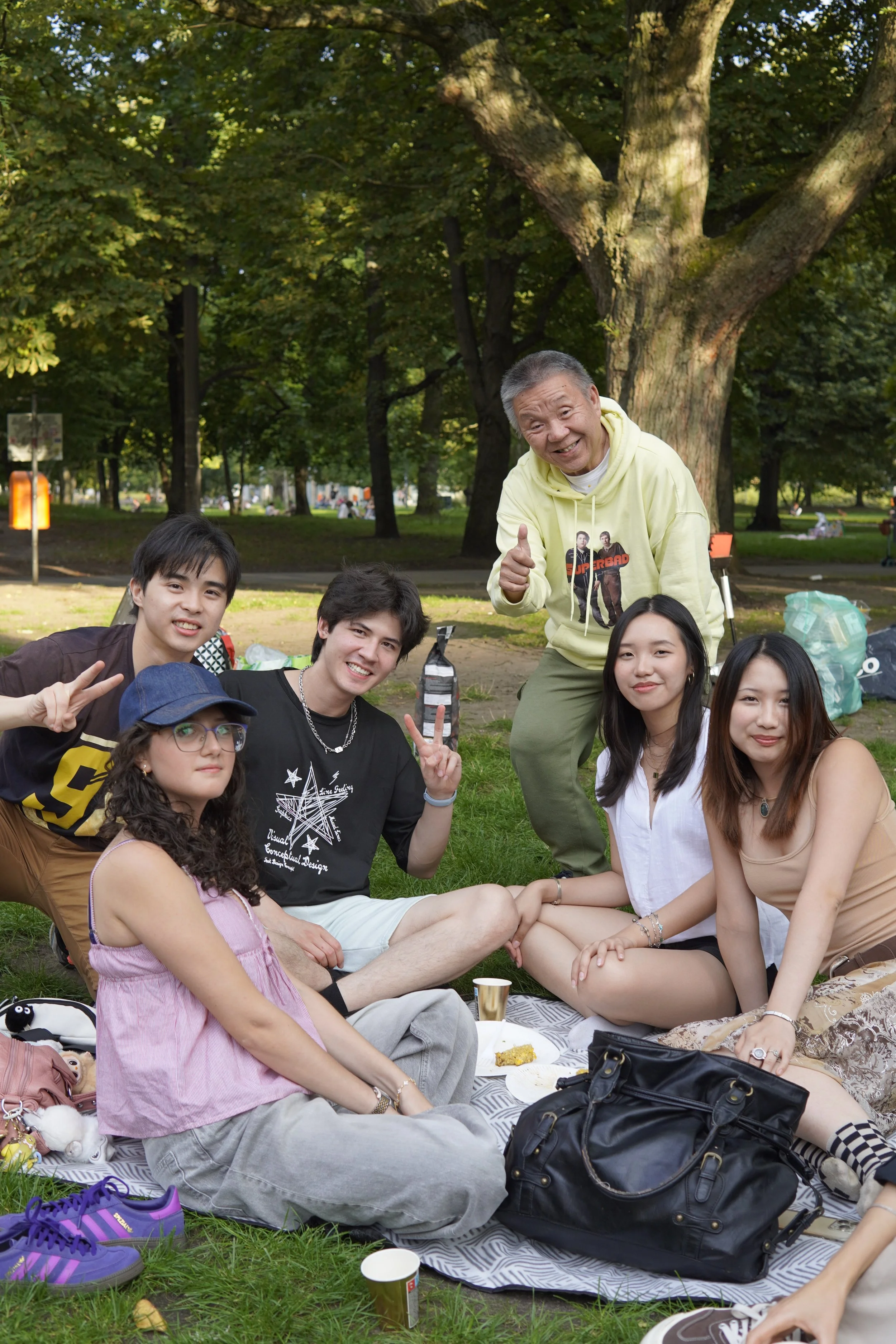 A group of five young people and one older man sitting on a picnic blanket in a park, smiling and making peace signs. They are surrounded by trees and outdoor equipment.