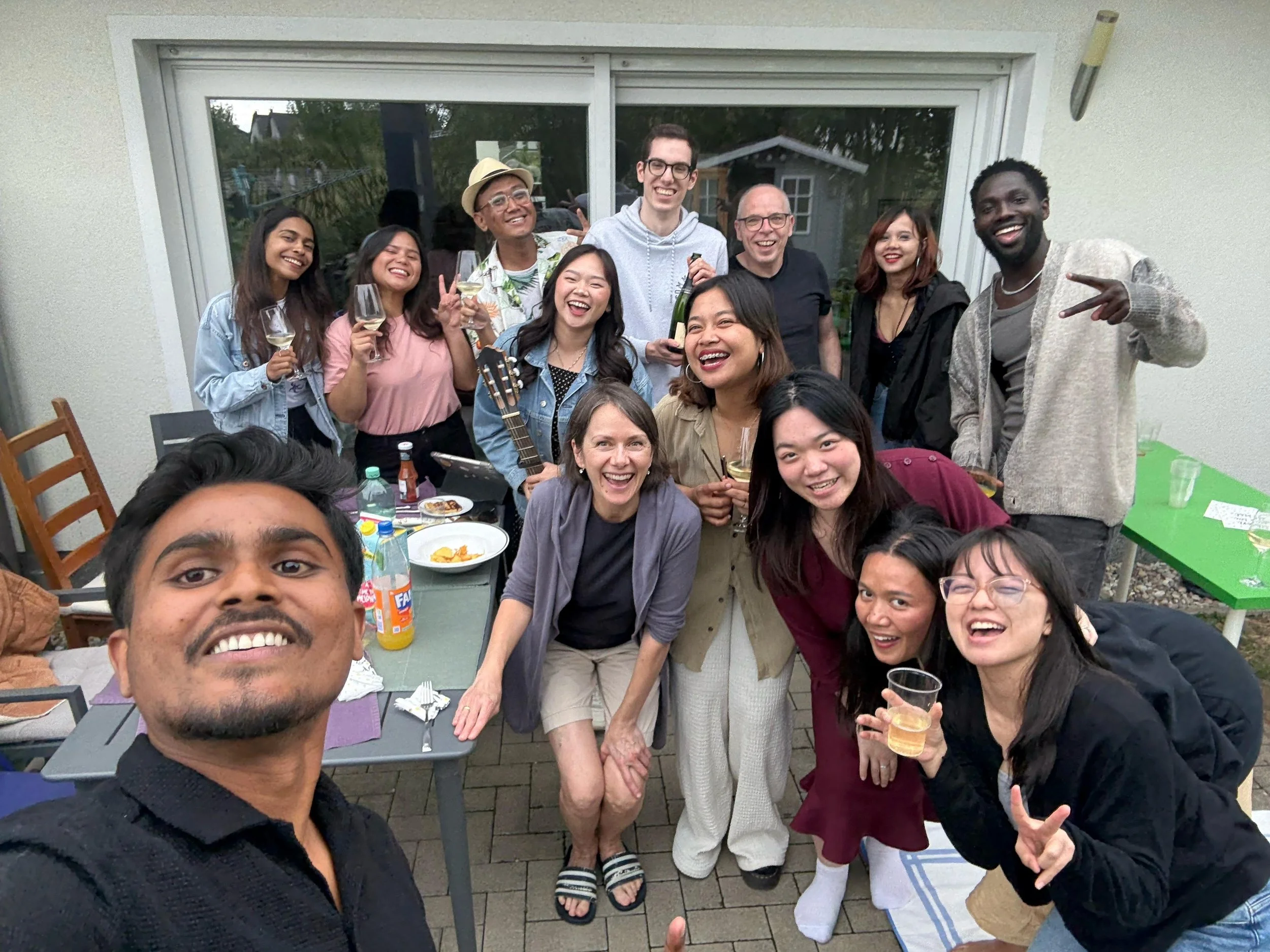 A diverse group of people at an outdoor gathering, smiling and holding drinks, with some making peace signs.