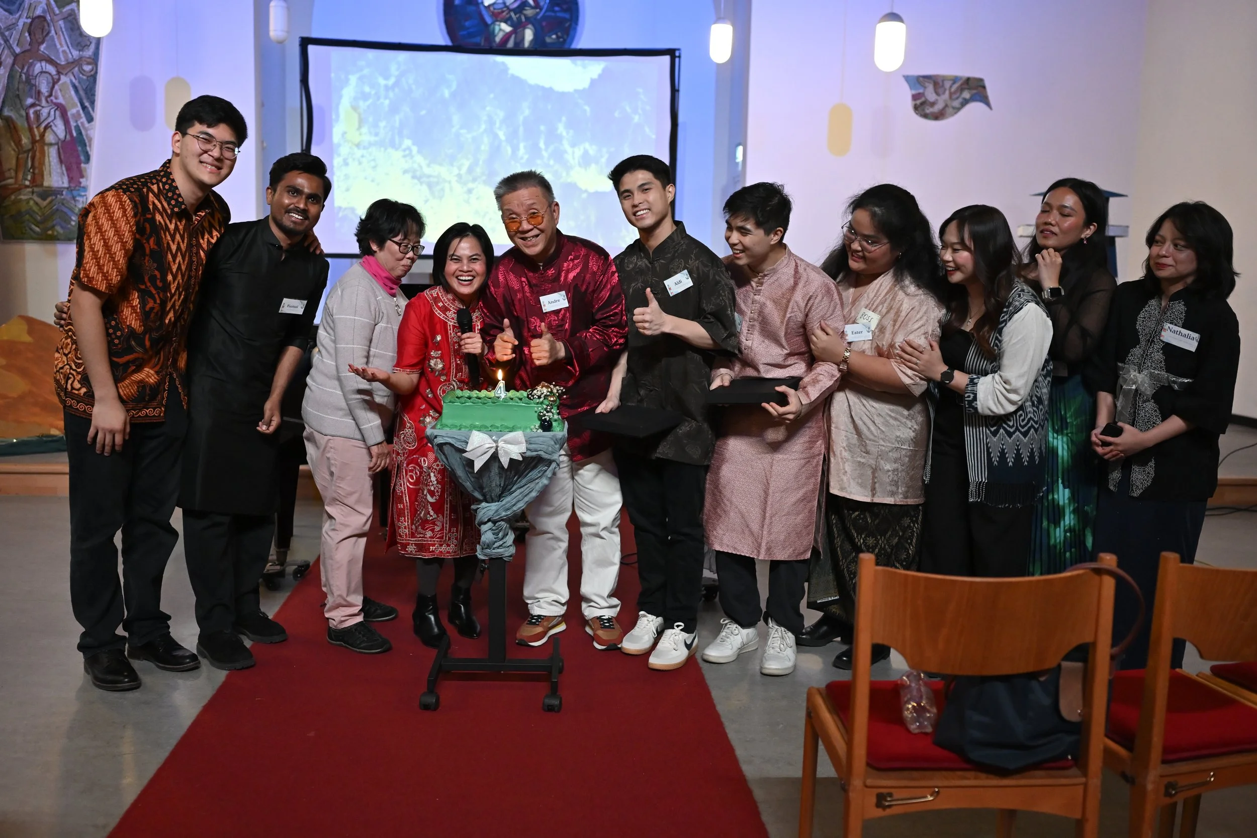 Group of people celebrating a birthday with a cake, some holding gift boxes, standing on a red carpet in a decorated indoor setting.