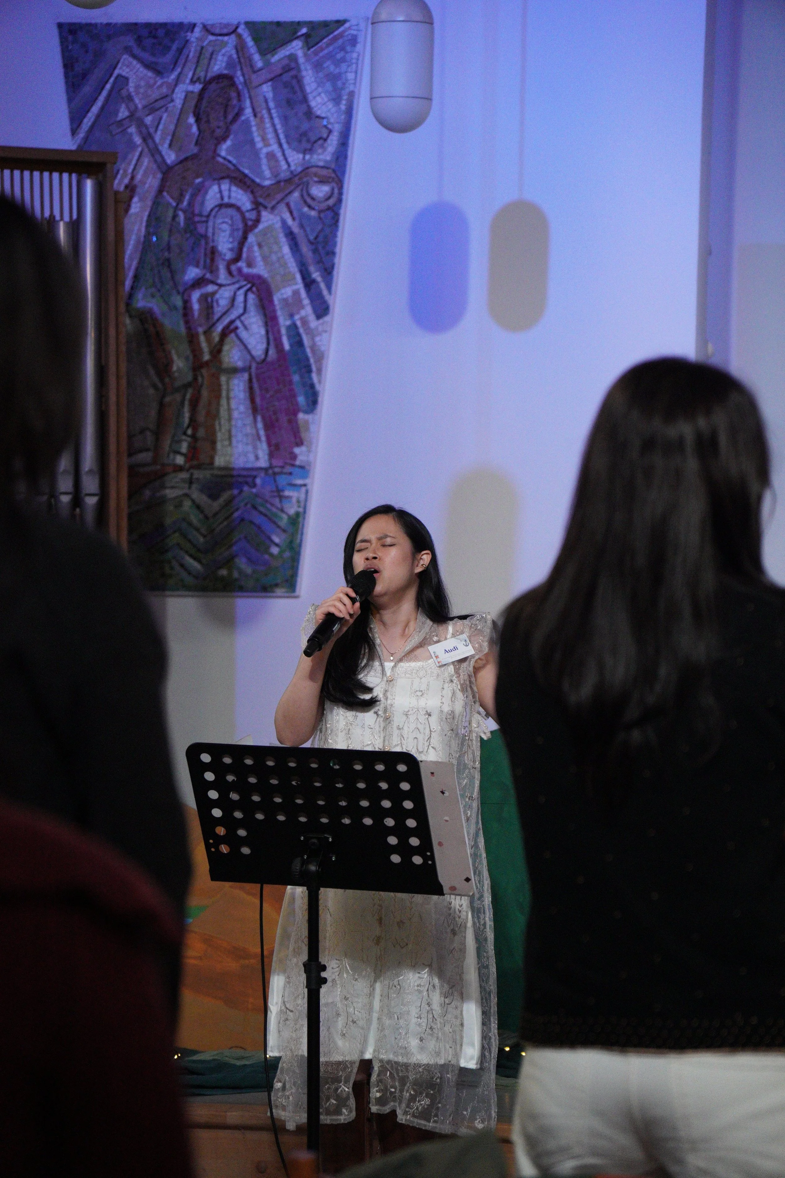A woman singing into a microphone during a performance, standing behind a music stand, with an audience in front of her. A religious-style artwork is hanging on the wall behind her.