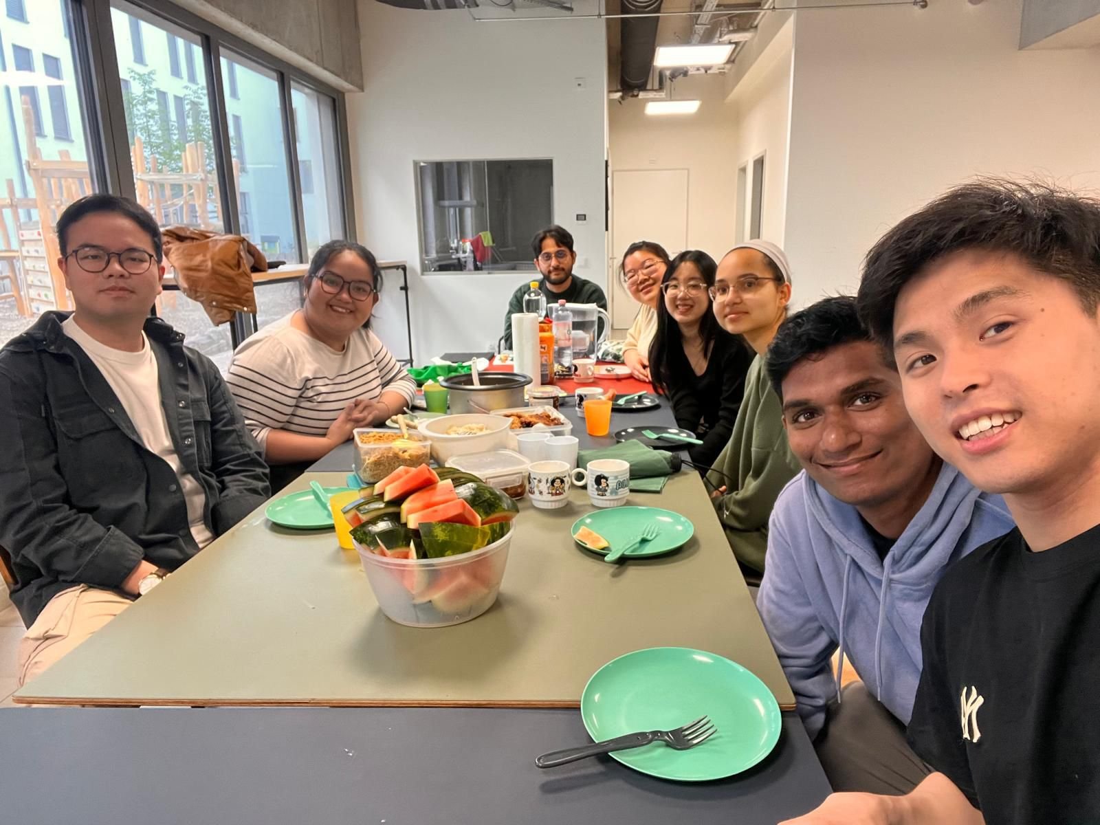 A group of nine diverse young adults sitting around a rectangular table in a modern apartment, celebrating with food and drinks, with a large bowl of watermelon slices on the table and various dishes and beverages.