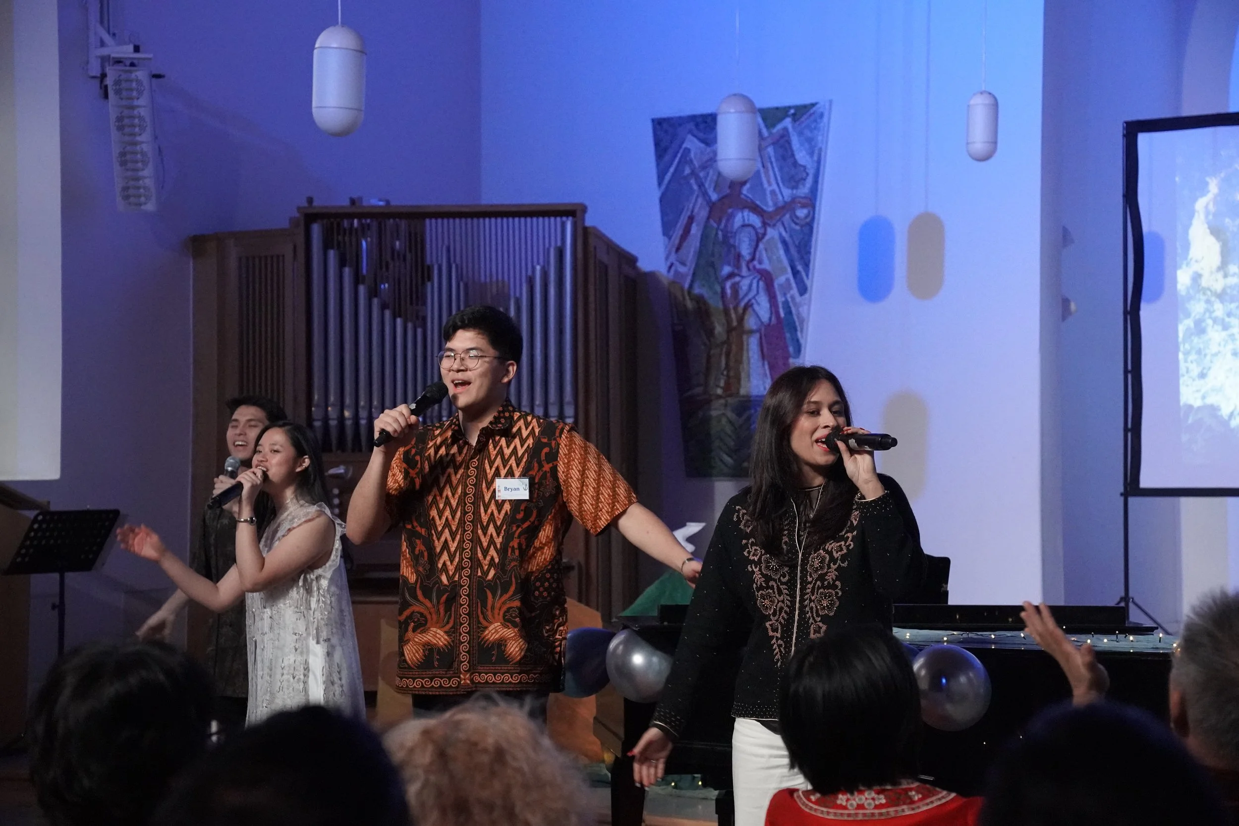 Group of four people singing on stage with microphones, audience in foreground, piano decorated with balloons, church window, and colorful artwork in background.