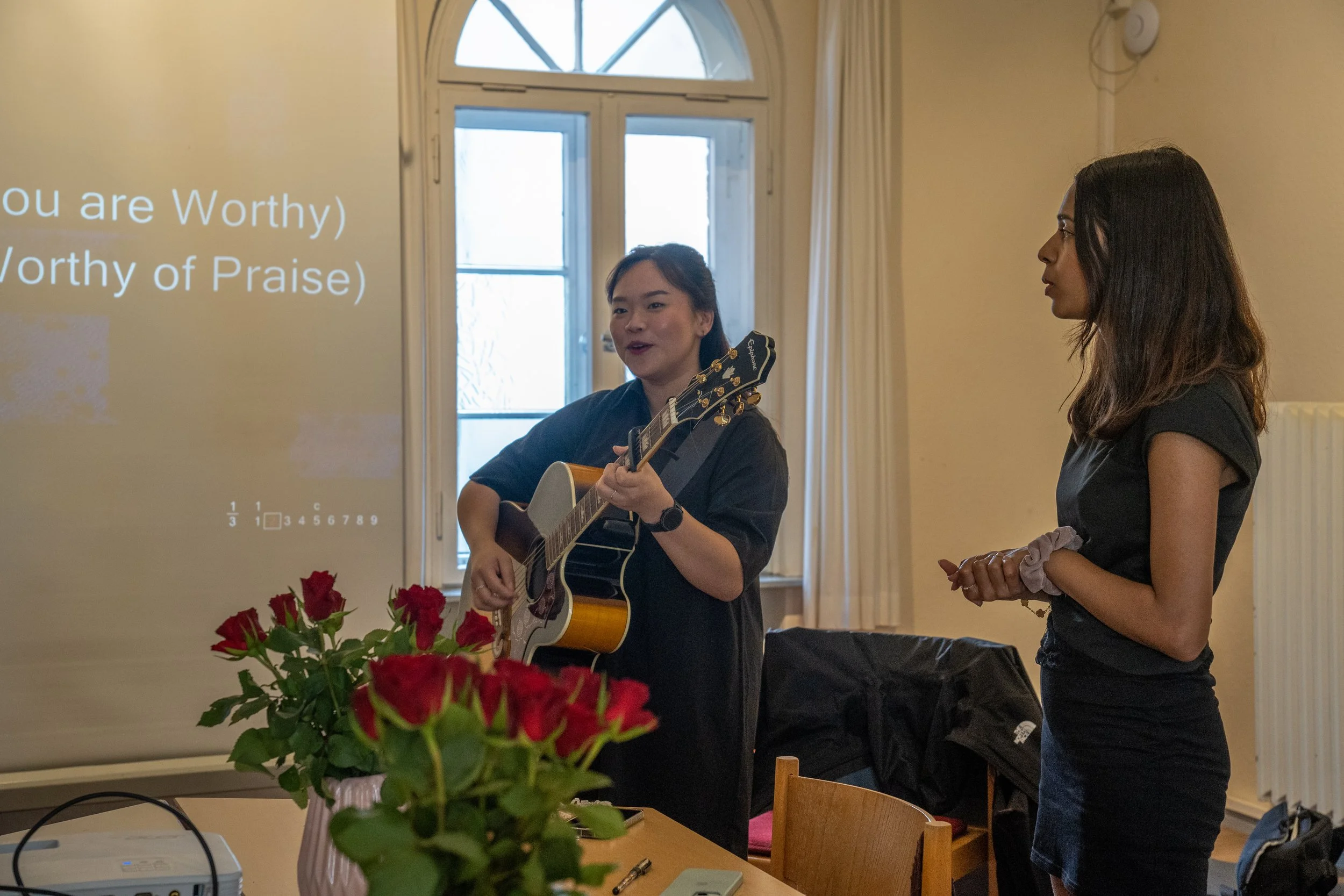 Two women stand in a room near a window, one playing an acoustic guitar and singing, the other observing. A vase of red roses is on the table in front of them, and a projector is on the table. A projection with text, 'You are Worthy, Worthy of Praise