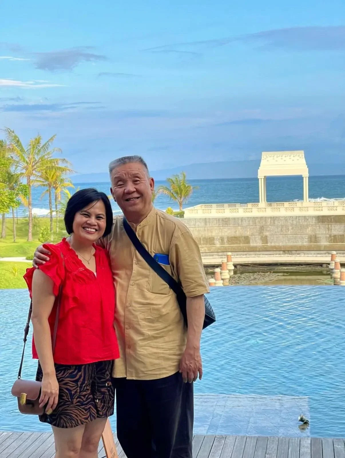 A smiling couple standing outdoors near a pool with the ocean and blue sky in the background, palm trees, and a white architectural structure.