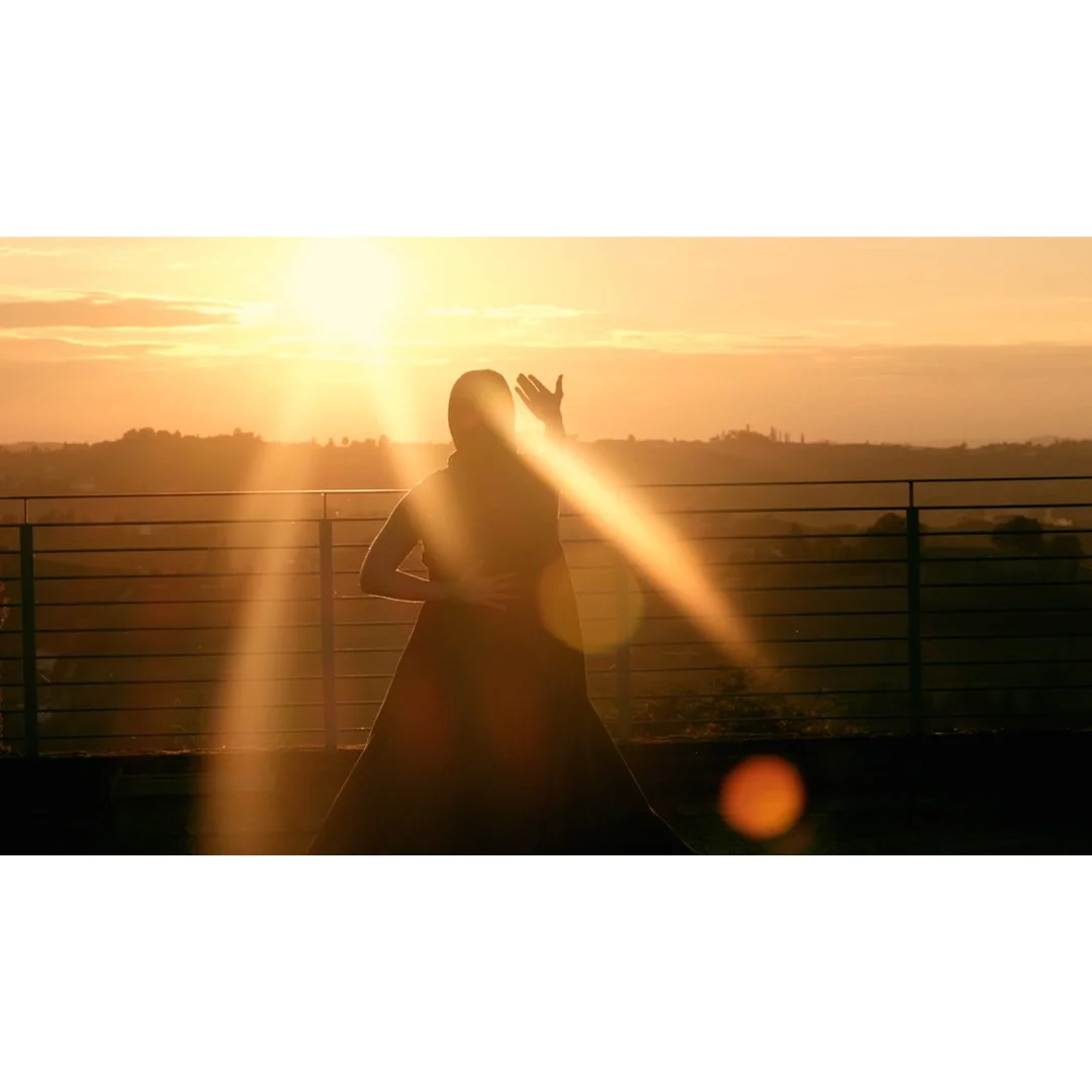 Figura di una donna silhouette che danza o si muove sulla terrazza durante il tramonto, con il sole e un cielo arancione sullo sfondo.