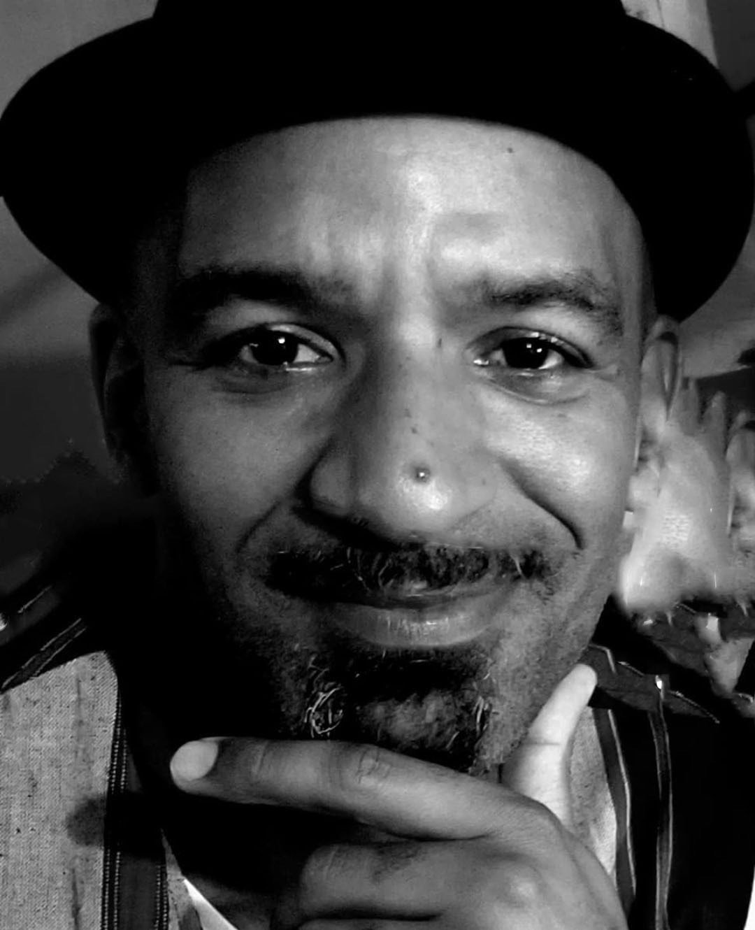 A close-up black and white photo of a man wearing a hat, smiling with his index finger on his chin, and sporting a small nose piercing.