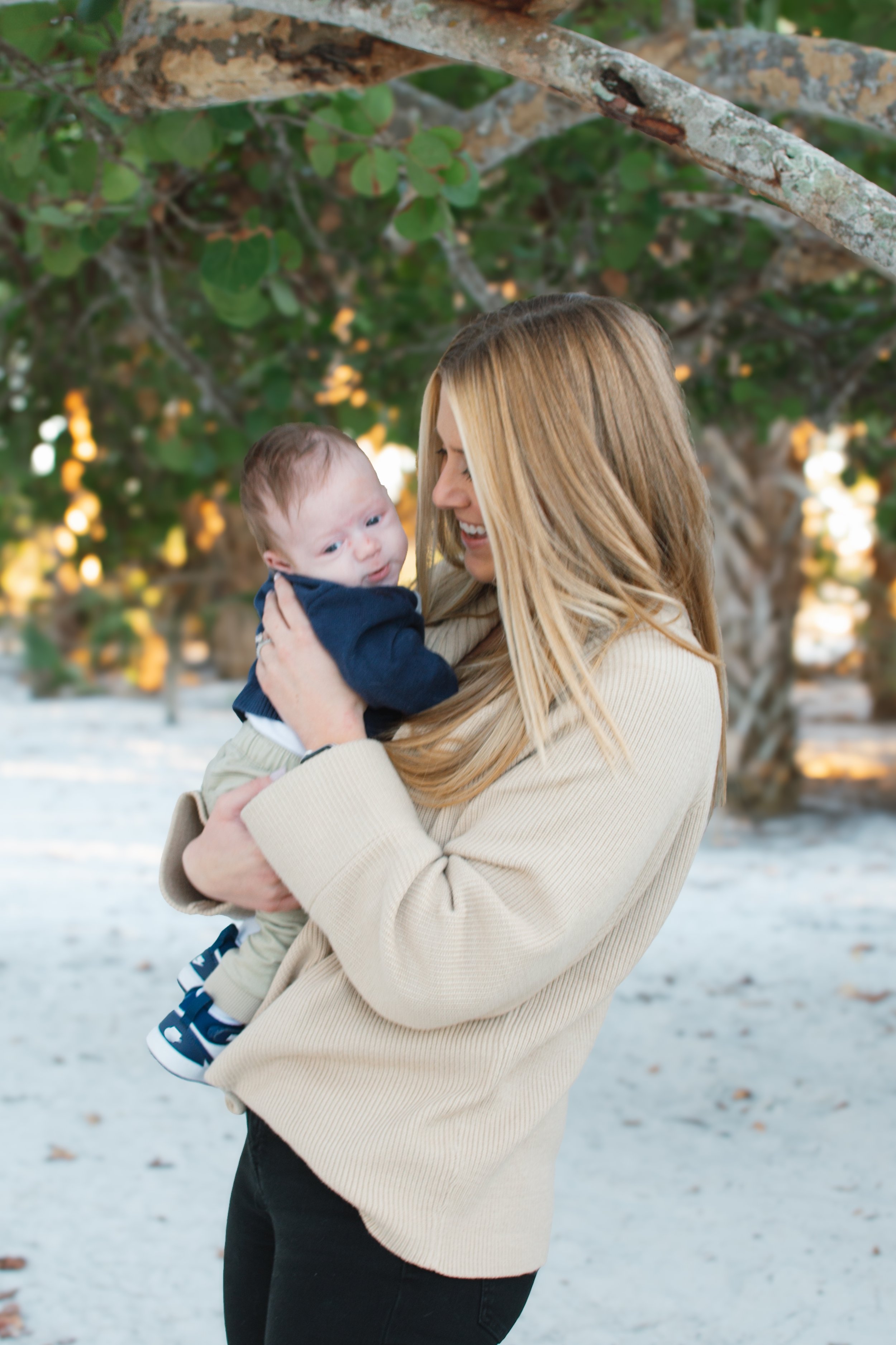 A woman with long blonde hair holding a baby boy outdoors under tree branches, smiling and looking at each other.
