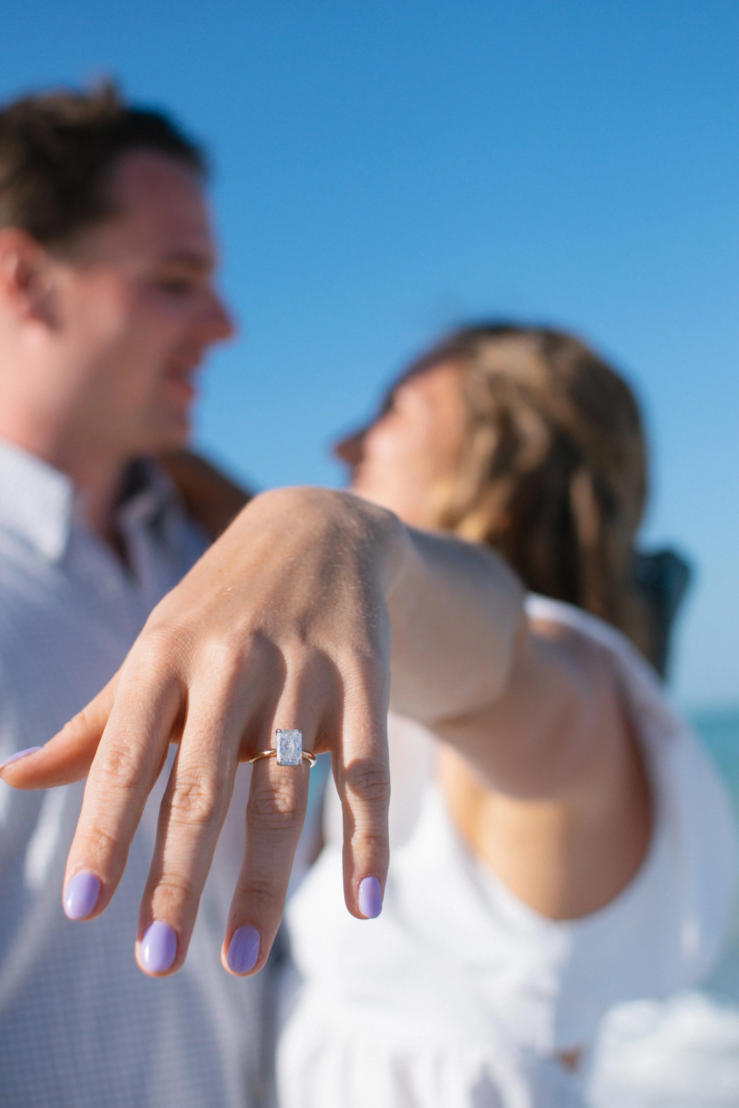 A woman displays a large rectangular-cut diamond engagement ring on her finger as a man and woman embrace in the background at the beach on a sunny day.