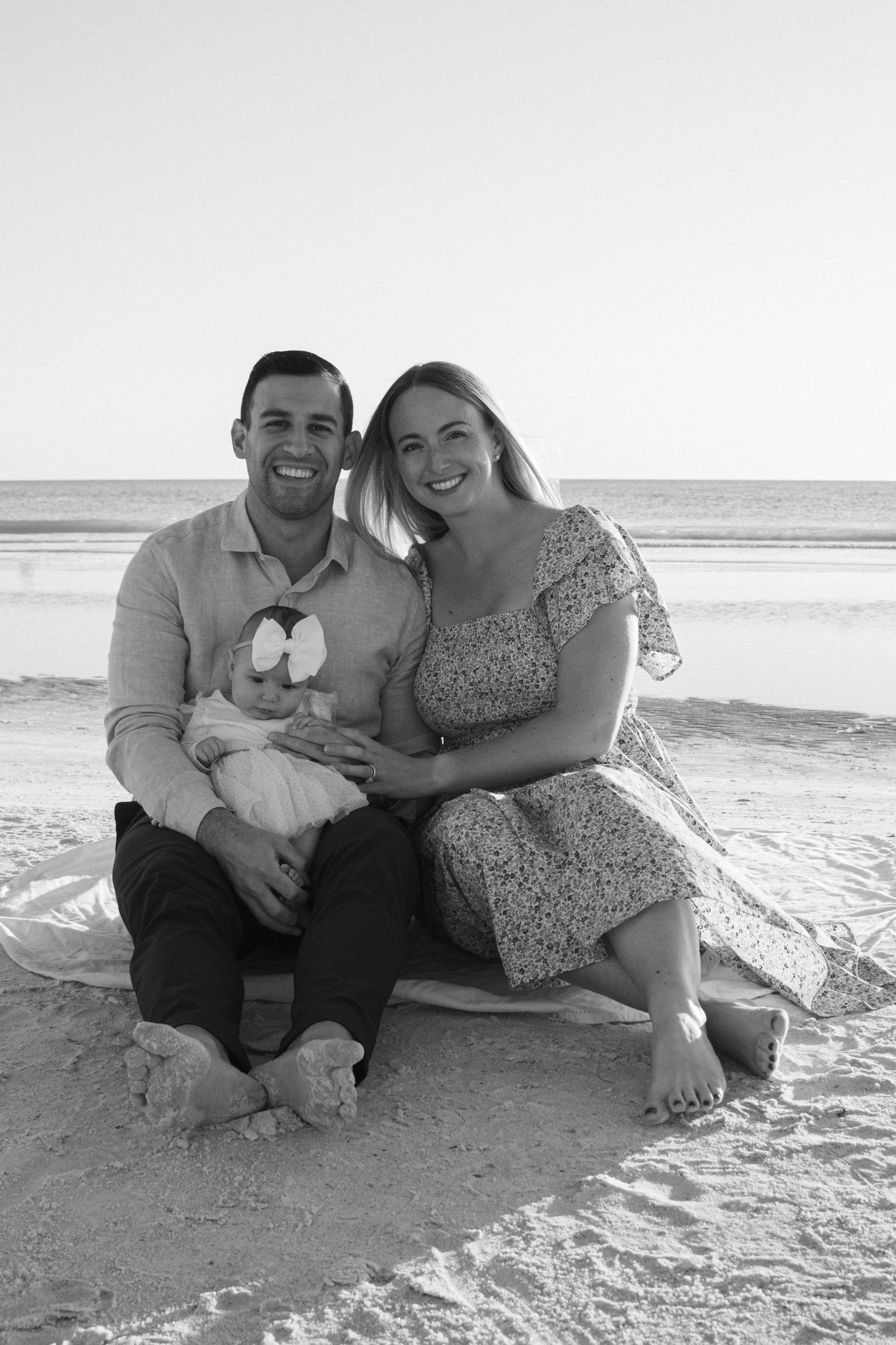 A happy family of three, including a man, woman, and baby girl, sitting on the sand at the beach during sunset, smiling at the camera.