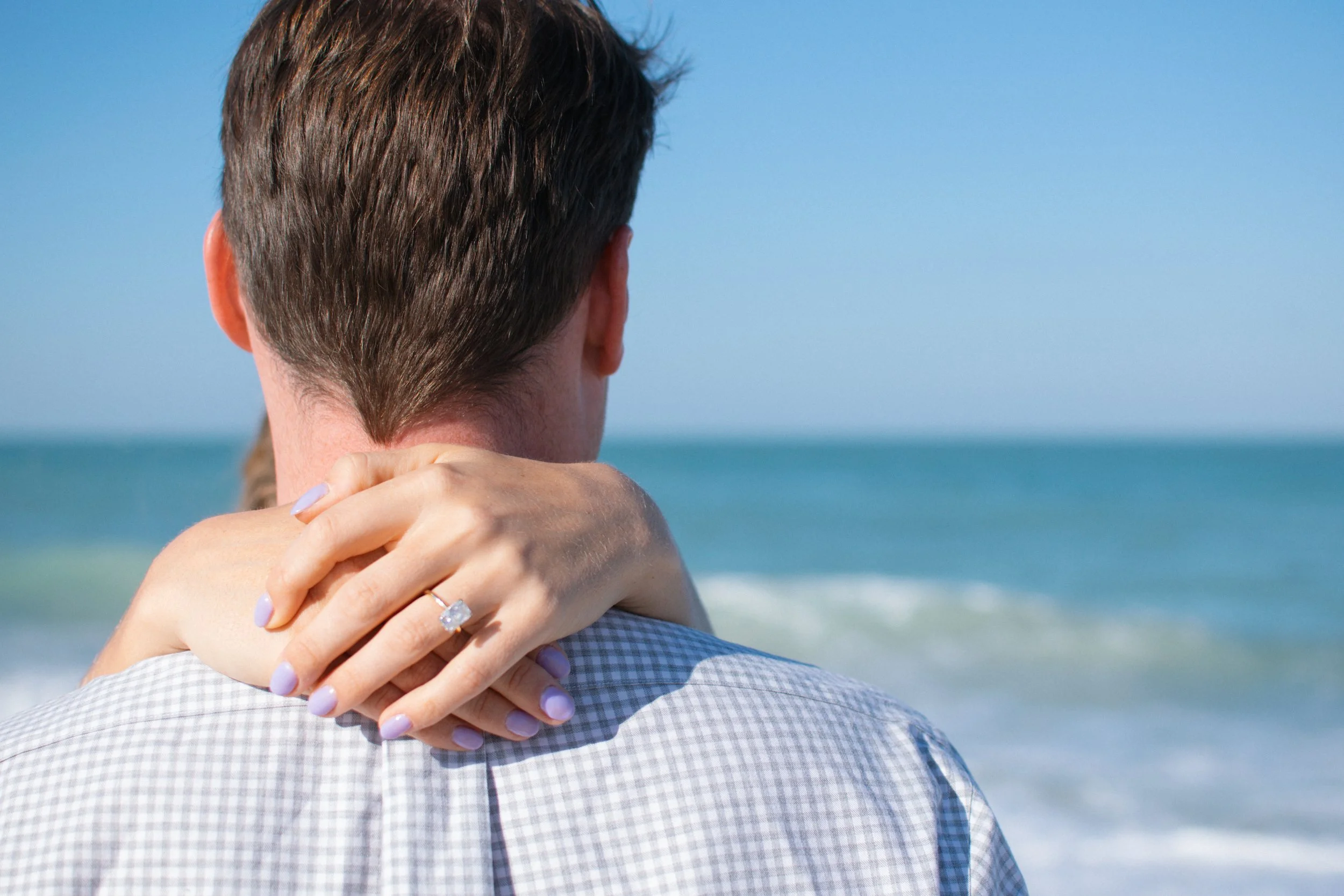 A woman hugging a man from behind on the beach with the ocean in the background, showing her hand with an engagement ring