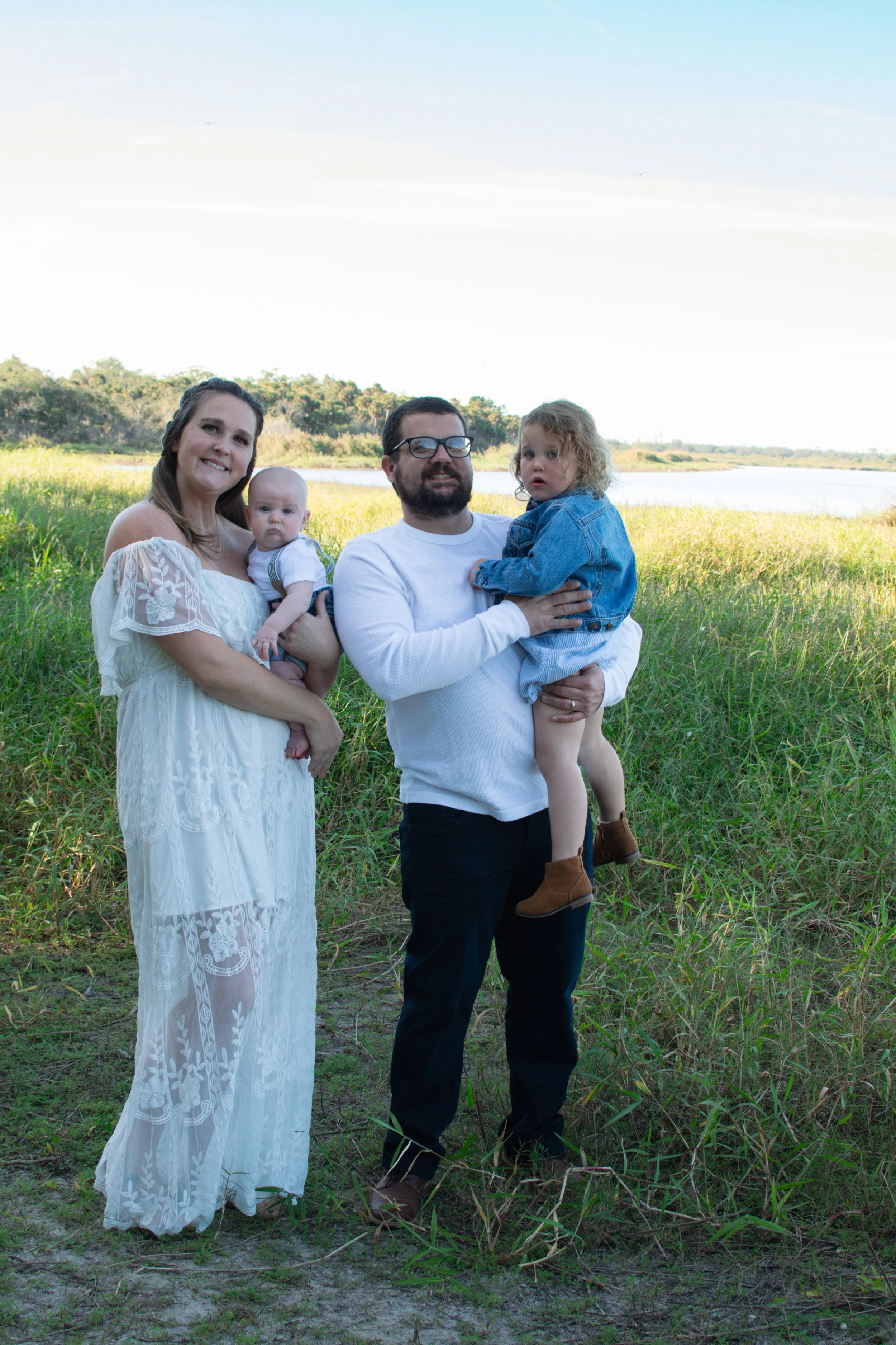A family of four outdoors near a river, with green grass and trees in the background. The woman wears a white dress, the man in a white shirt and black pants, and they hold two young children, one in a denim jacket and the other in a white shirt. The