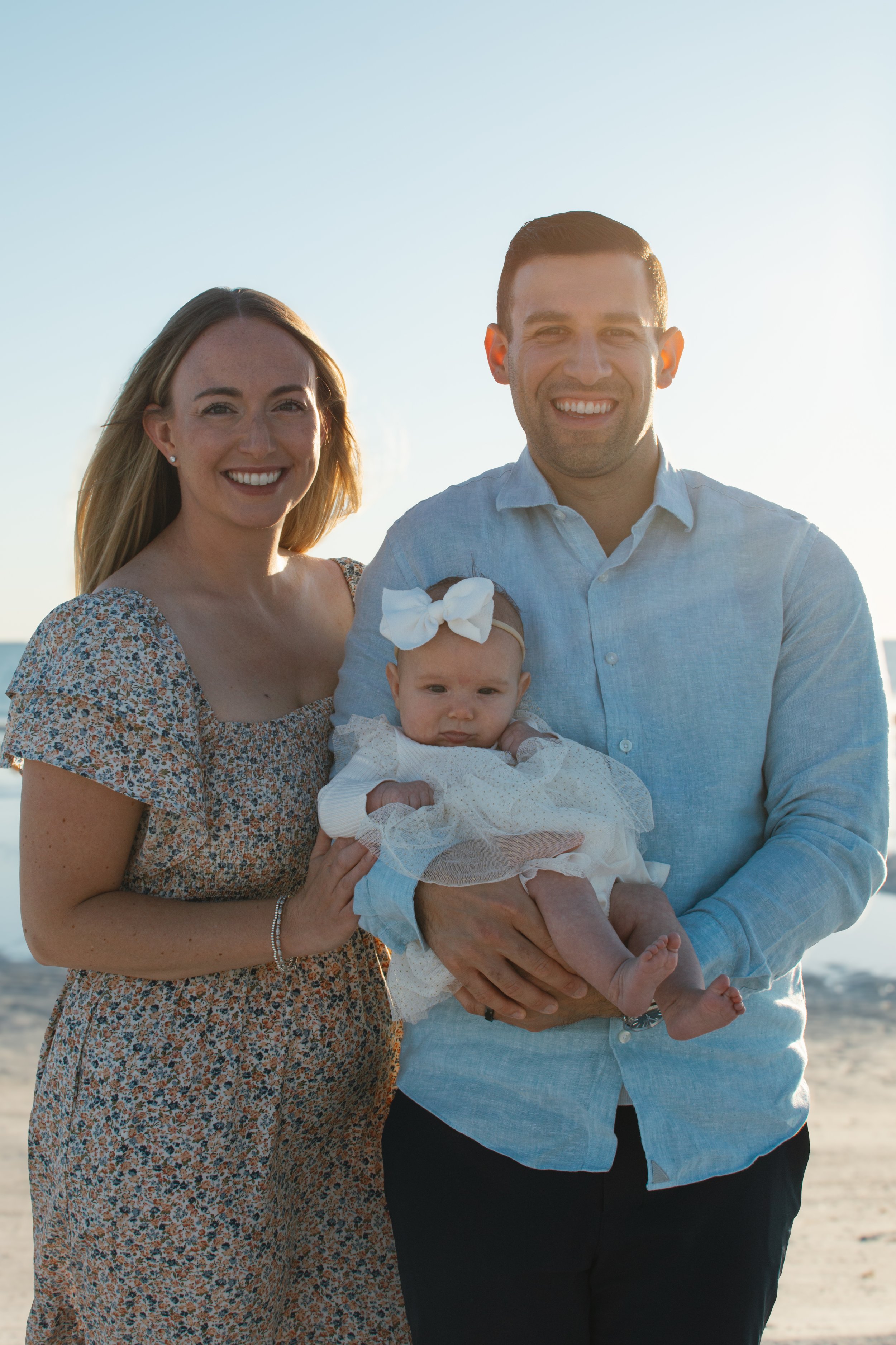 A happy family of three standing outdoors on a beach at sunset, holding a baby girl dressed in white.