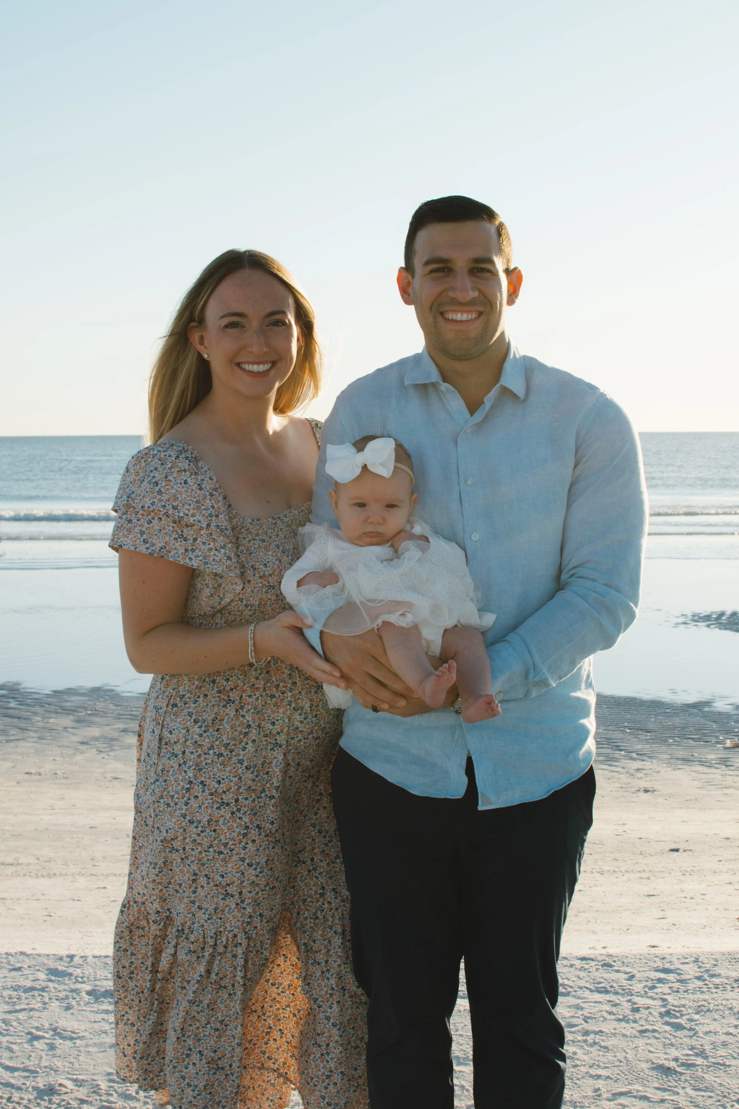 Family of three standing on a beach during sunset, woman, man, and baby girl in white dress with large bow, smiling at camera.
