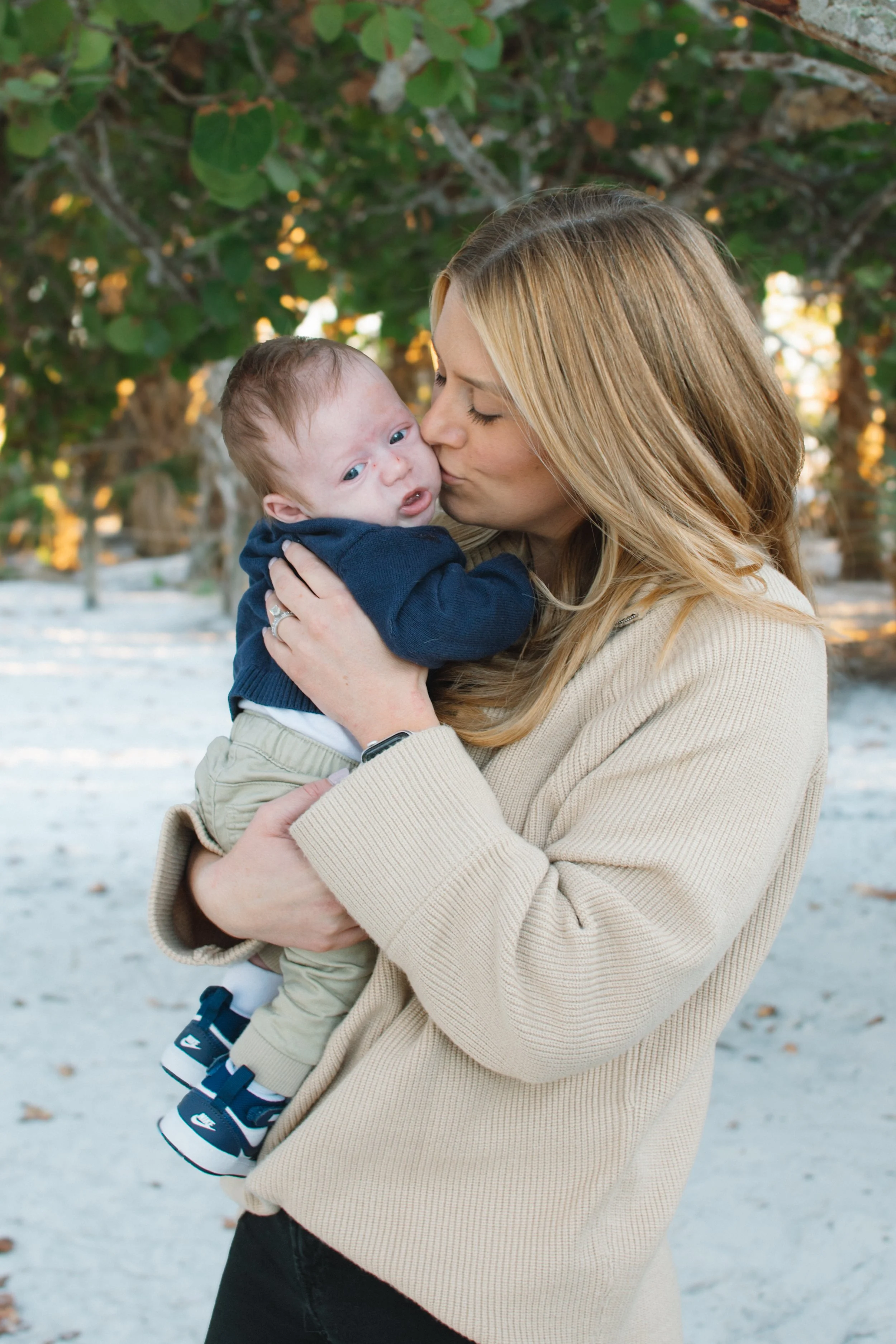 A woman is holding and kissing a baby outdoors near trees, with a sandy or snow-covered ground in the background.
