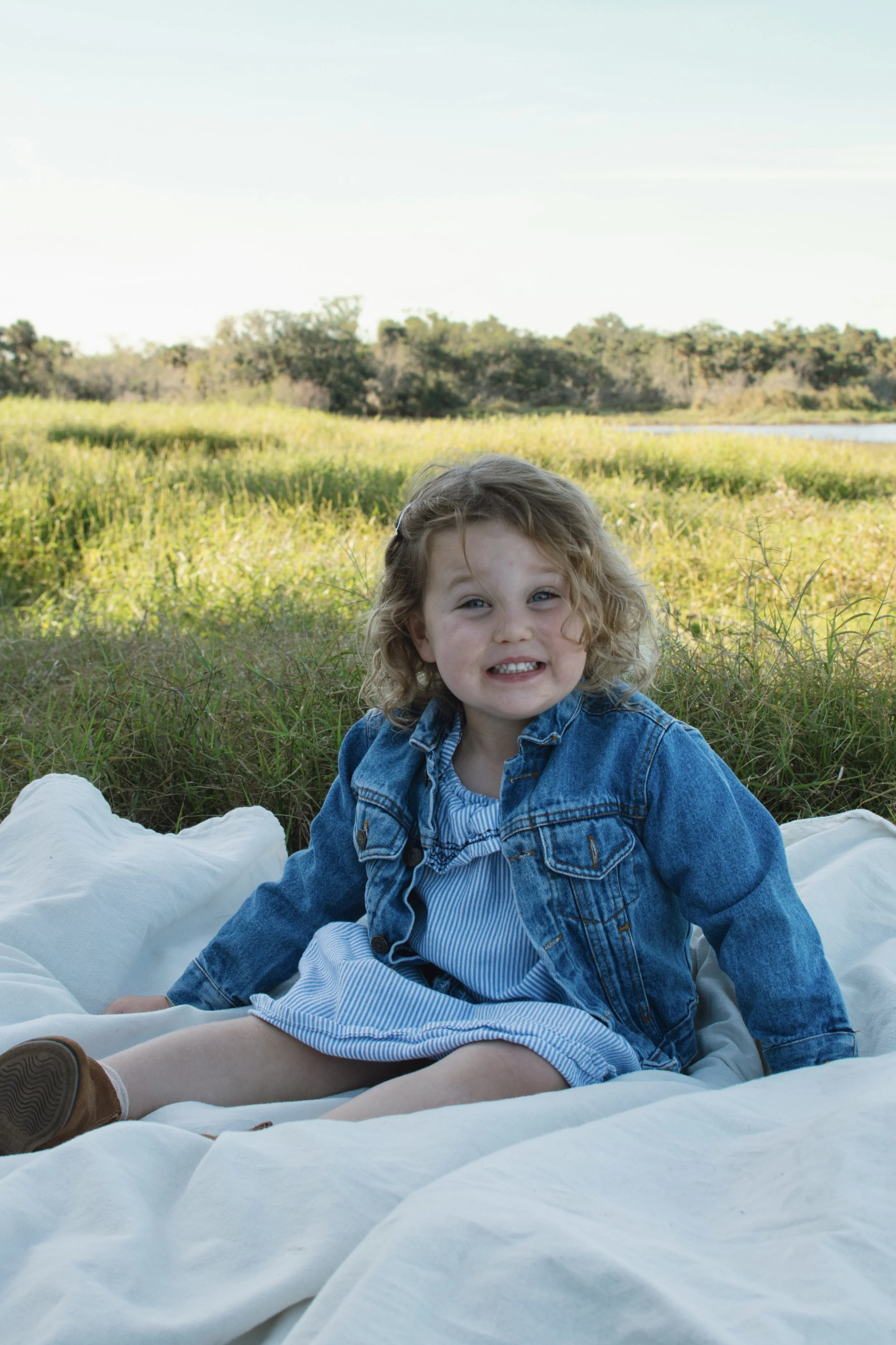 A young girl with curly blonde hair wearing a denim jacket and blue dress sitting on a white blanket outdoors in a grassy field with trees in the background.