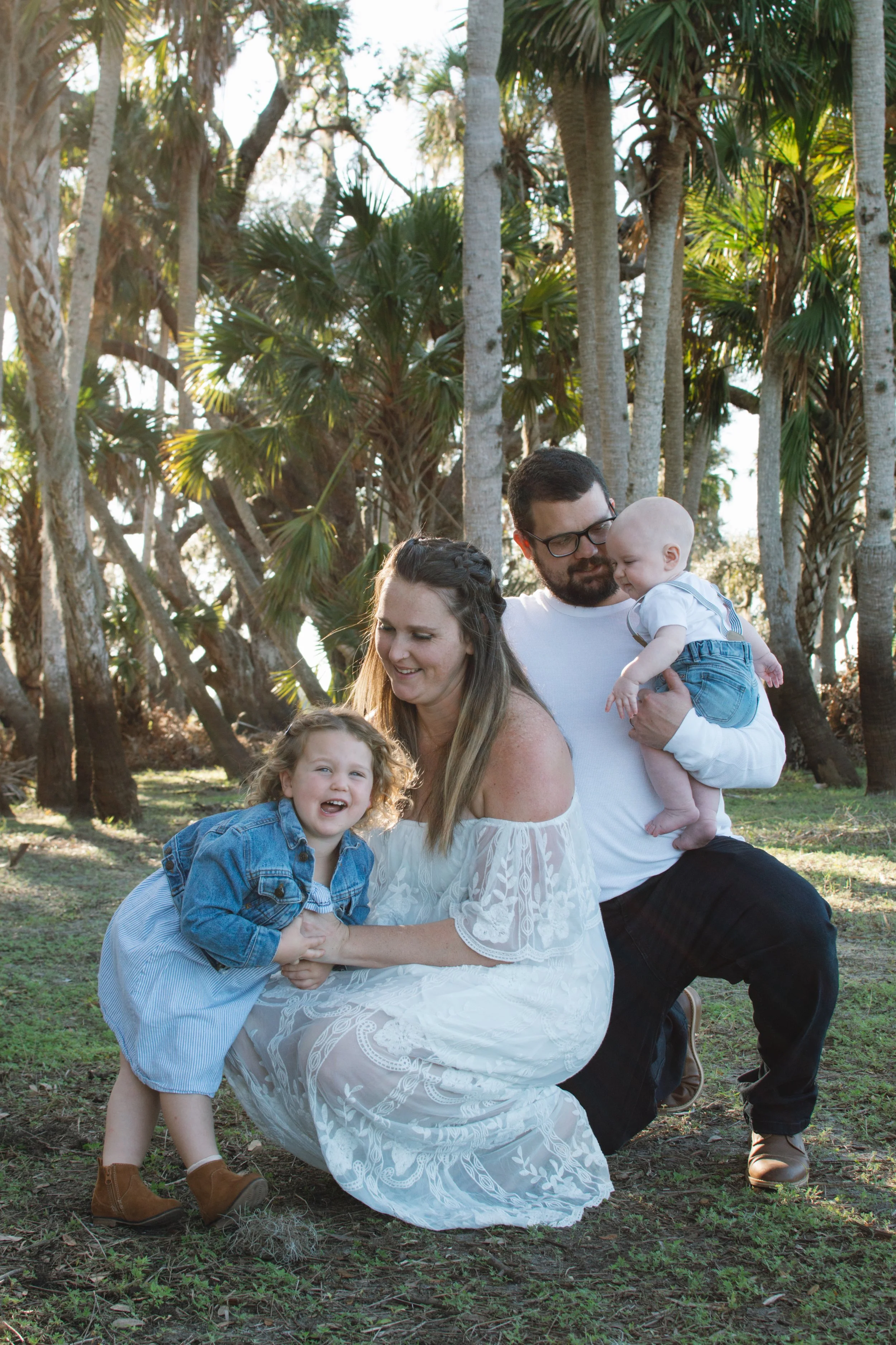 A family of four posing outdoors in a wooded area during daylight, with palm trees in the background. The mother is kneeling, wearing a white dress, smiling at her daughter. The father, holding a baby, is standing beside her. The daughter, dressed in