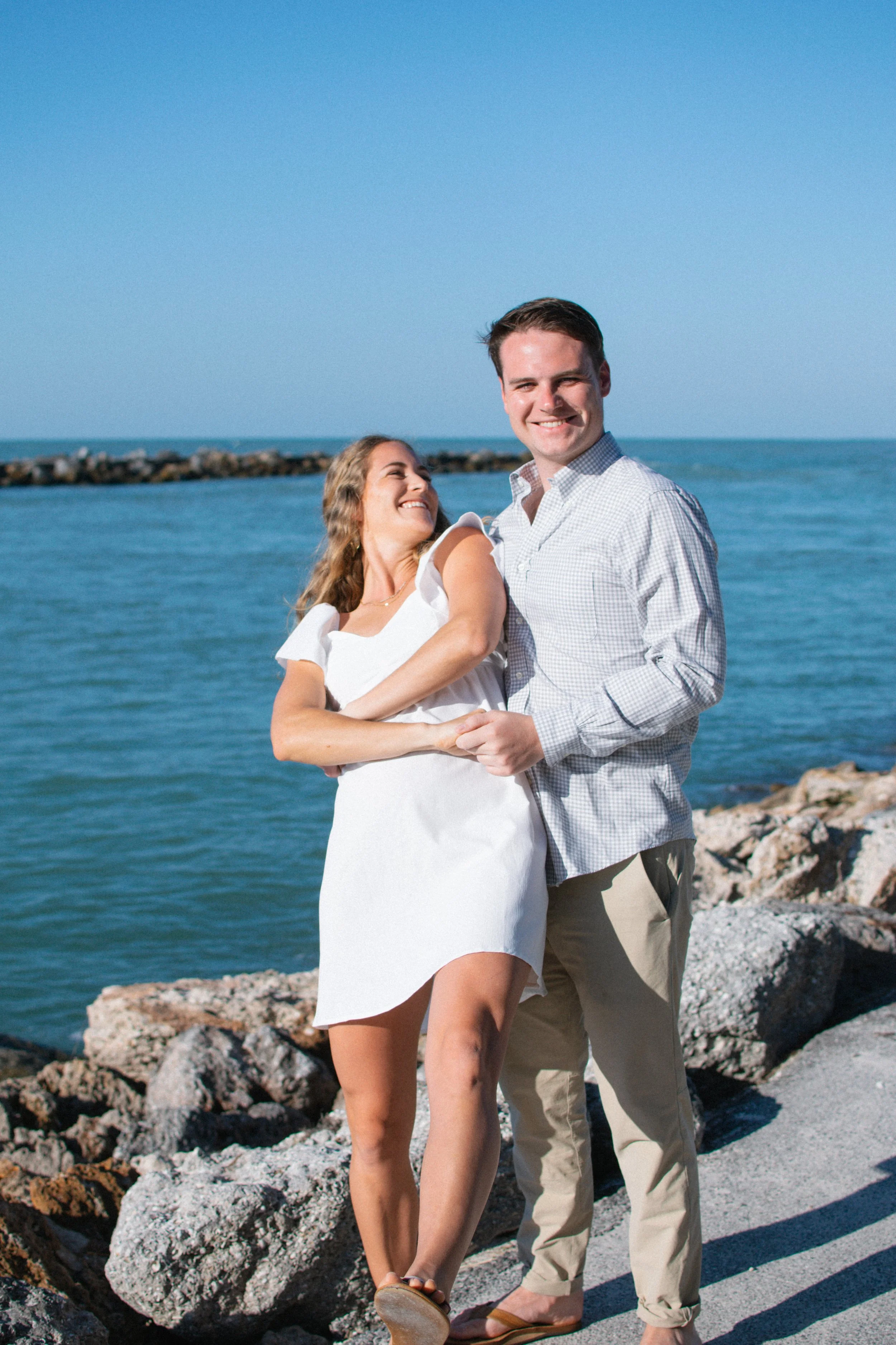 A smiling couple in casual clothing enjoying a walk along rocky shoreline by the ocean, with a breakwater in the background and clear blue sky.