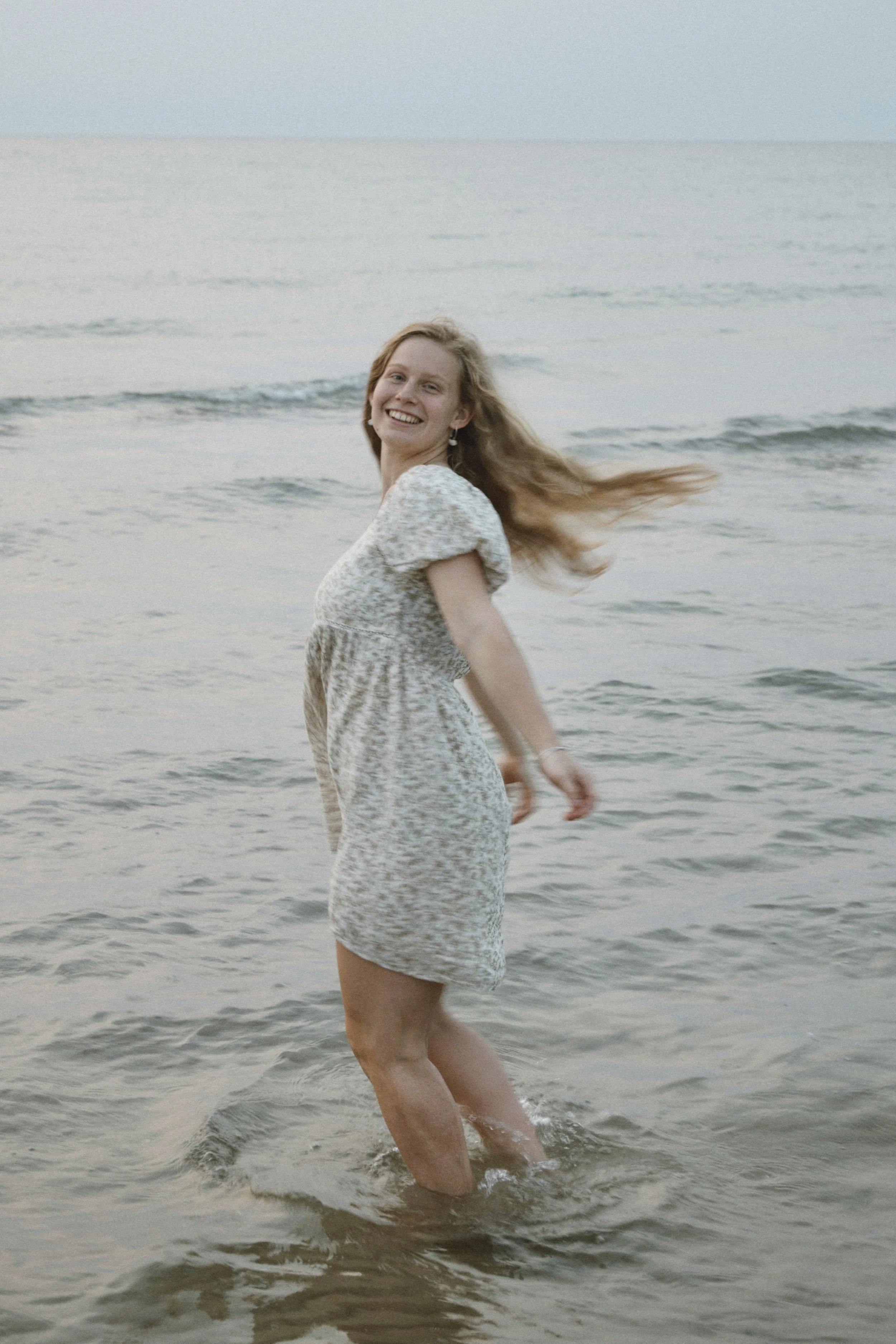A woman with long blonde hair smiling and spinning in shallow ocean water at the beach, wearing a light-colored, patterned dress.