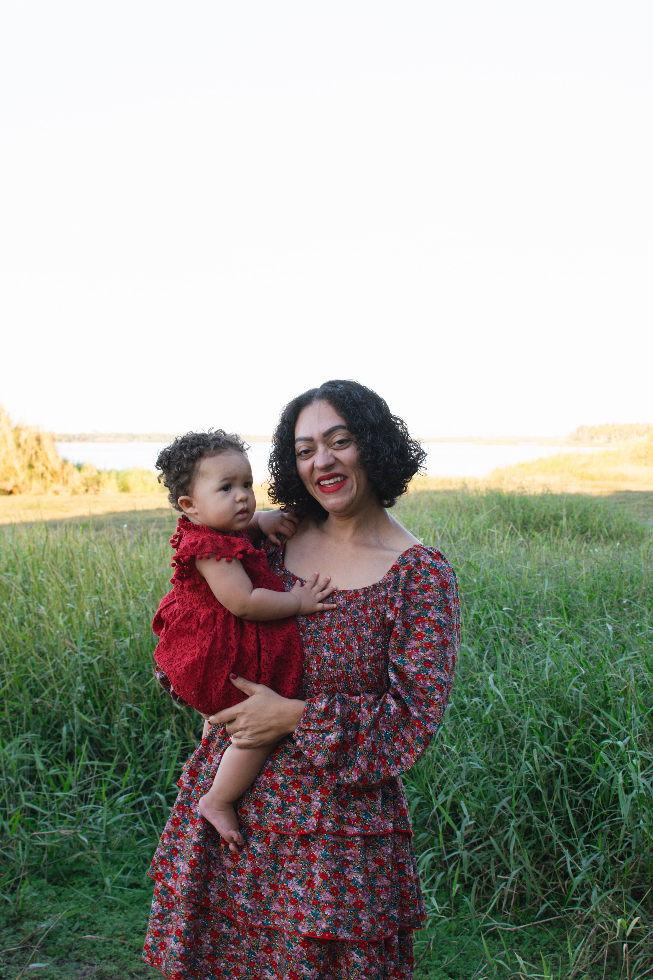 A woman with curly black hair and red lipstick holding a young girl with short curly hair in a red dress, standing in a grassy field with a body of water in the background on a clear day.