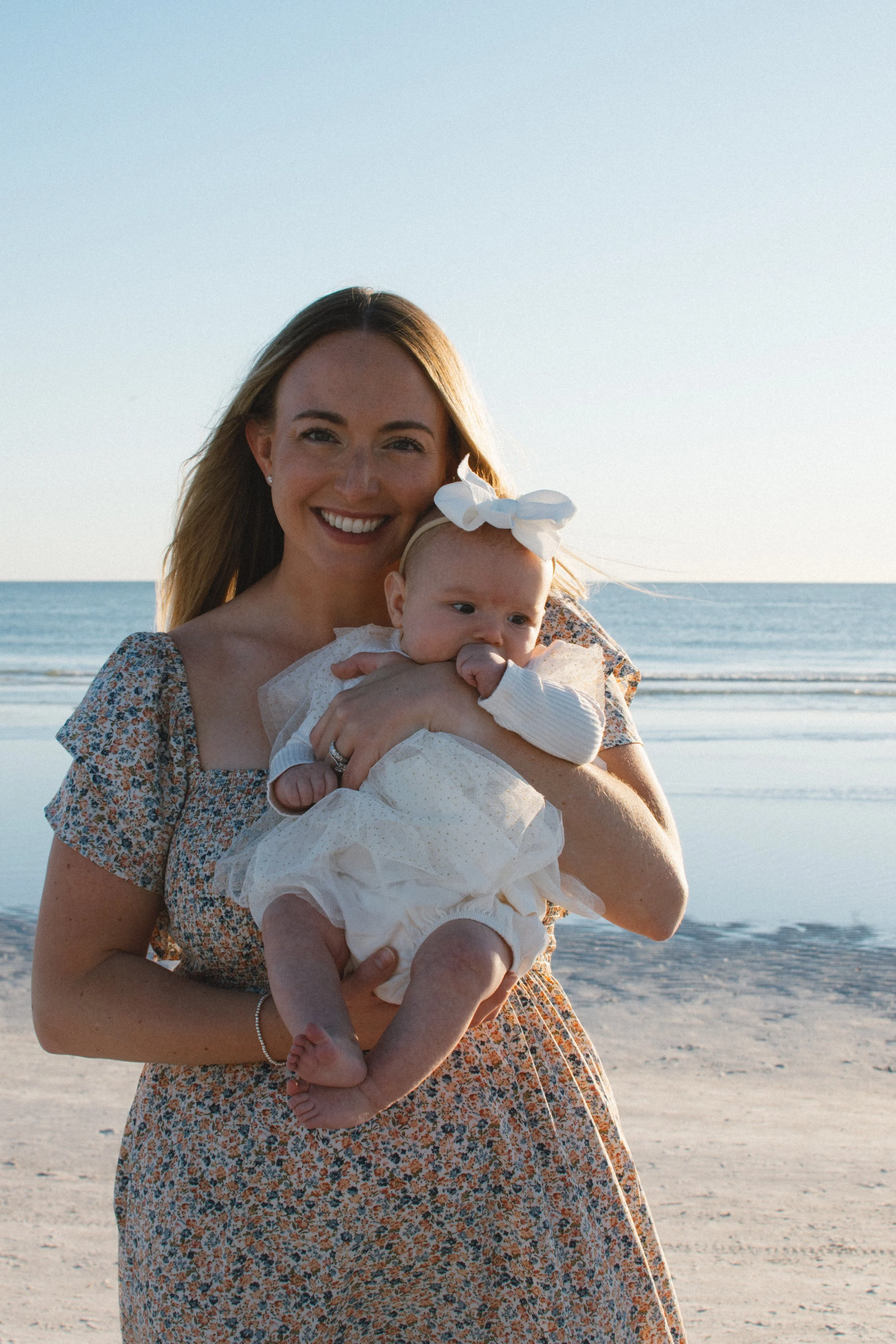 A young woman smiling and holding a baby girl on a beach with the ocean in the background.