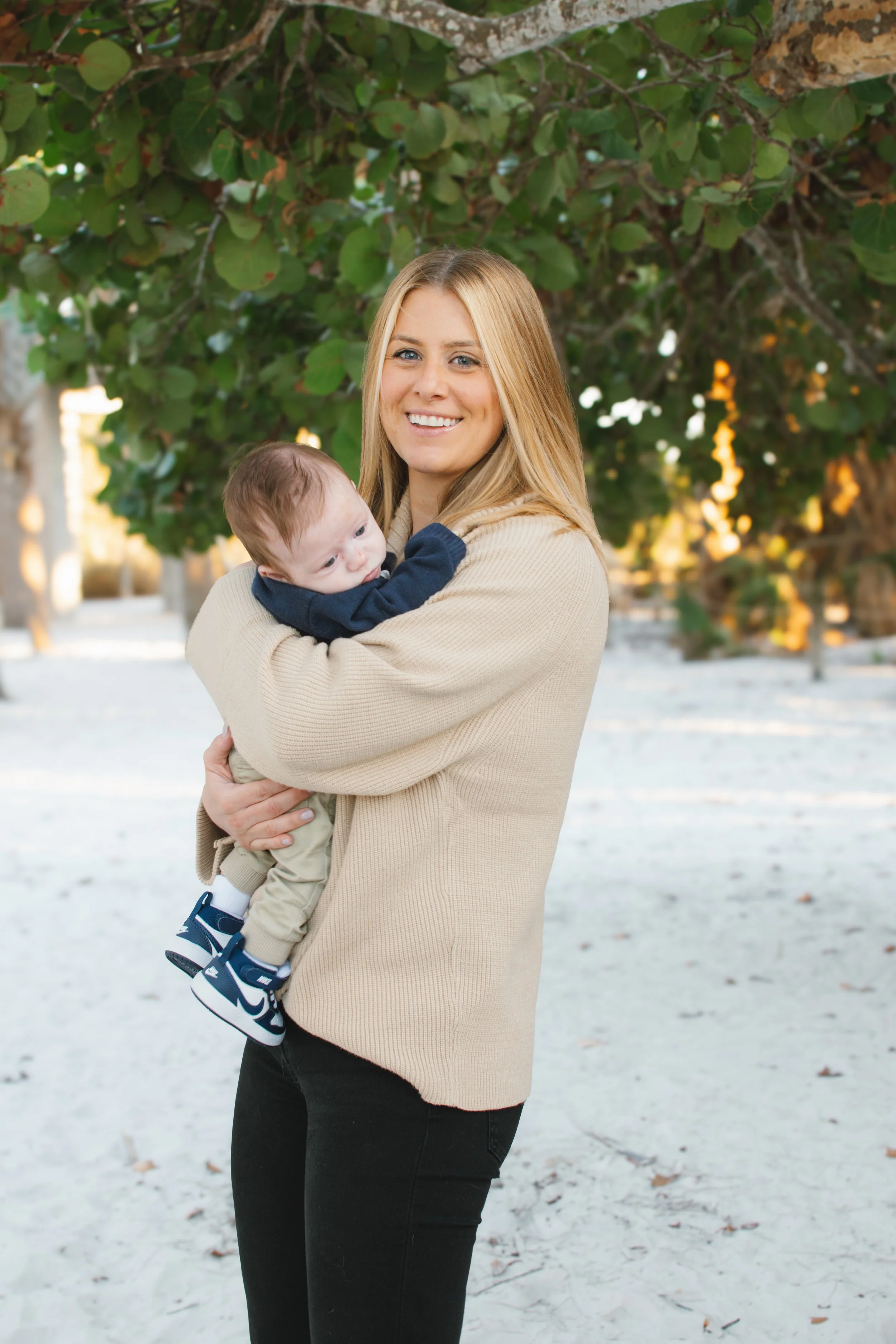 A woman holding a baby outdoors near a leafy tree with sunlight filtering through the branches.
