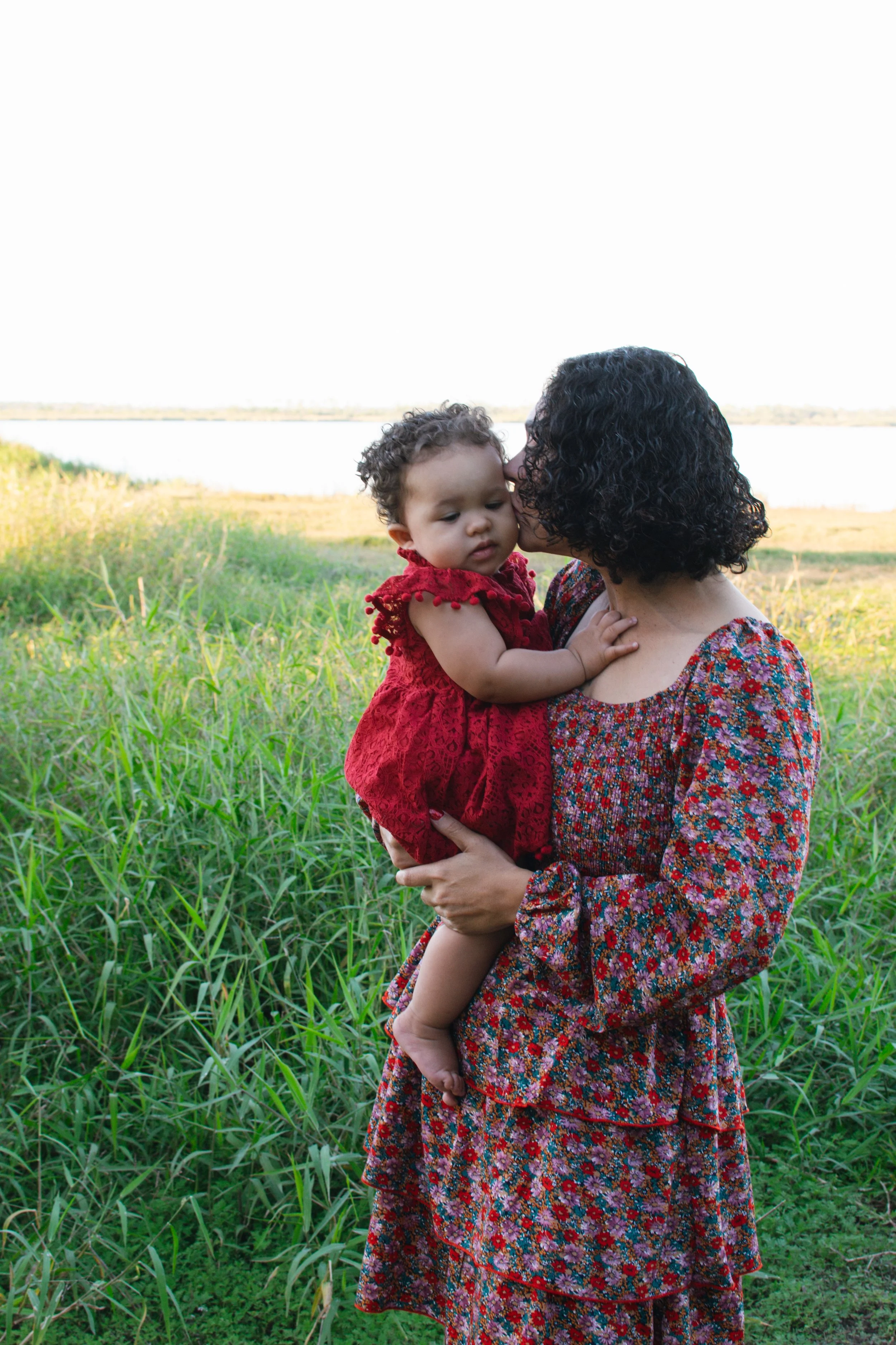 A woman with curly black hair holding a young child in a red dress near a water body, with tall grass in the foreground and a bright sky in the background.