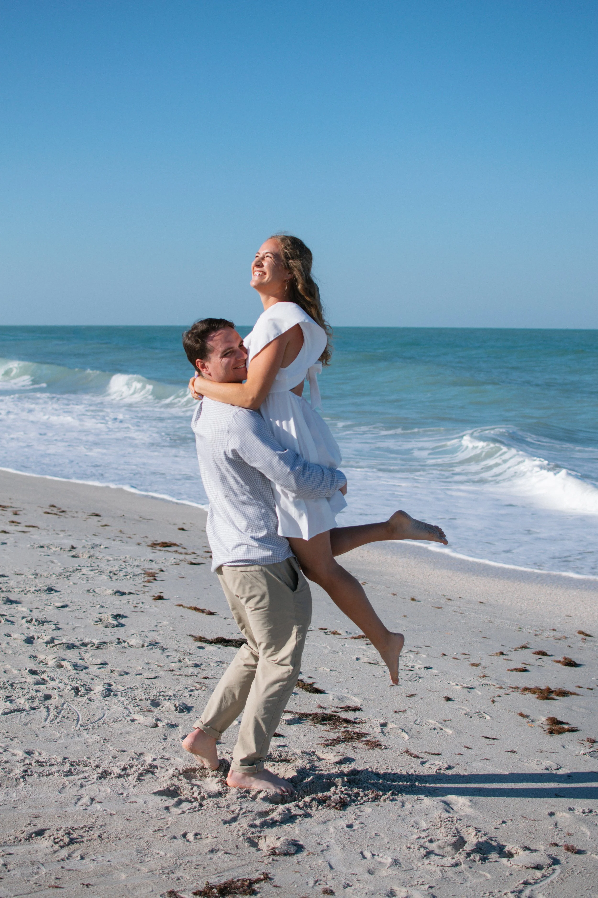 A joyful couple on the beach, with the man lifting the woman who is smiling with her eyes closed, near the shoreline with waves and a clear blue sky in the background.