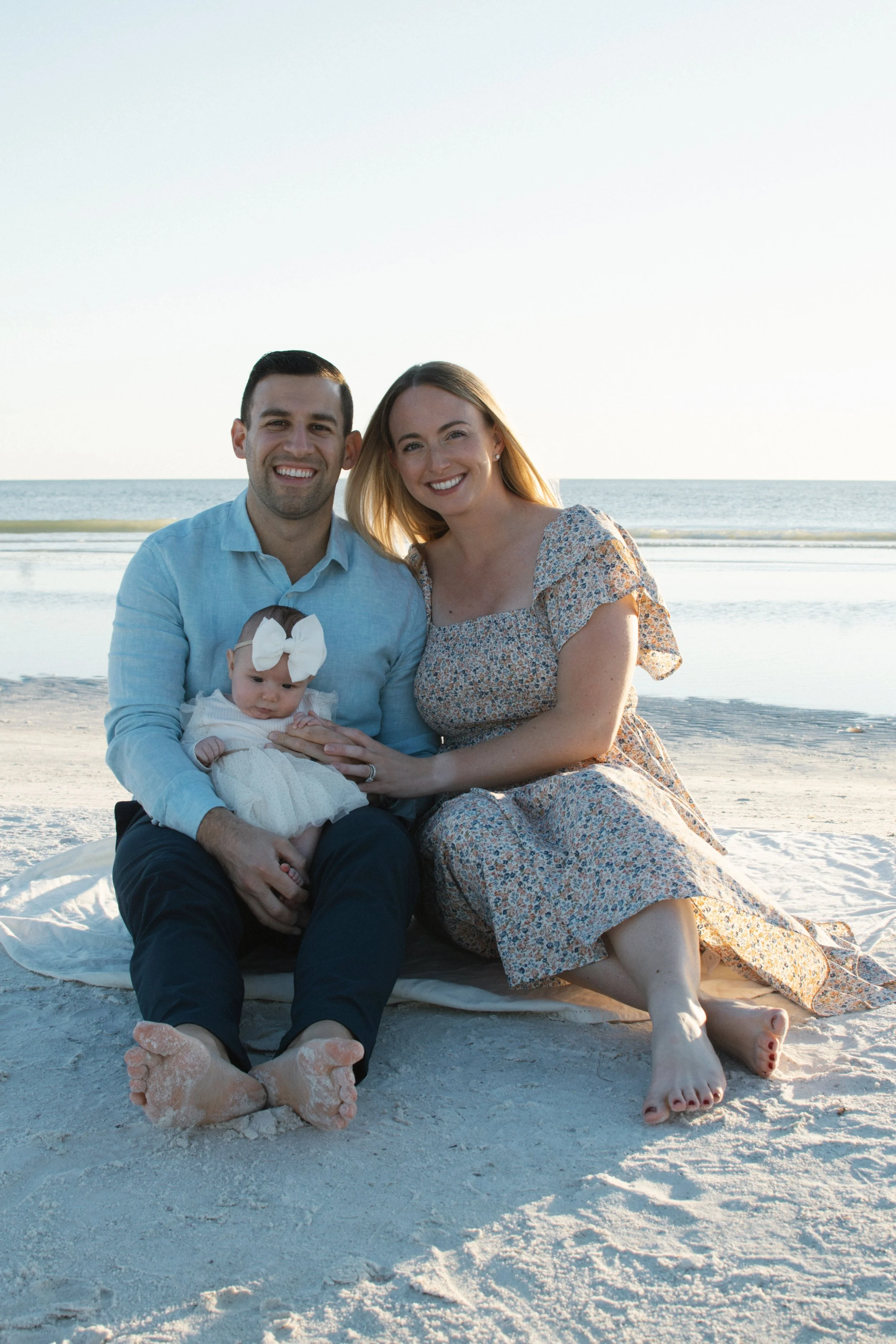 A family of three sitting on a sandy beach, smiling at the camera. The father, mother, and a baby girl with a large white bow on her head, dressed in a white dress. The ocean and sky are in the background.