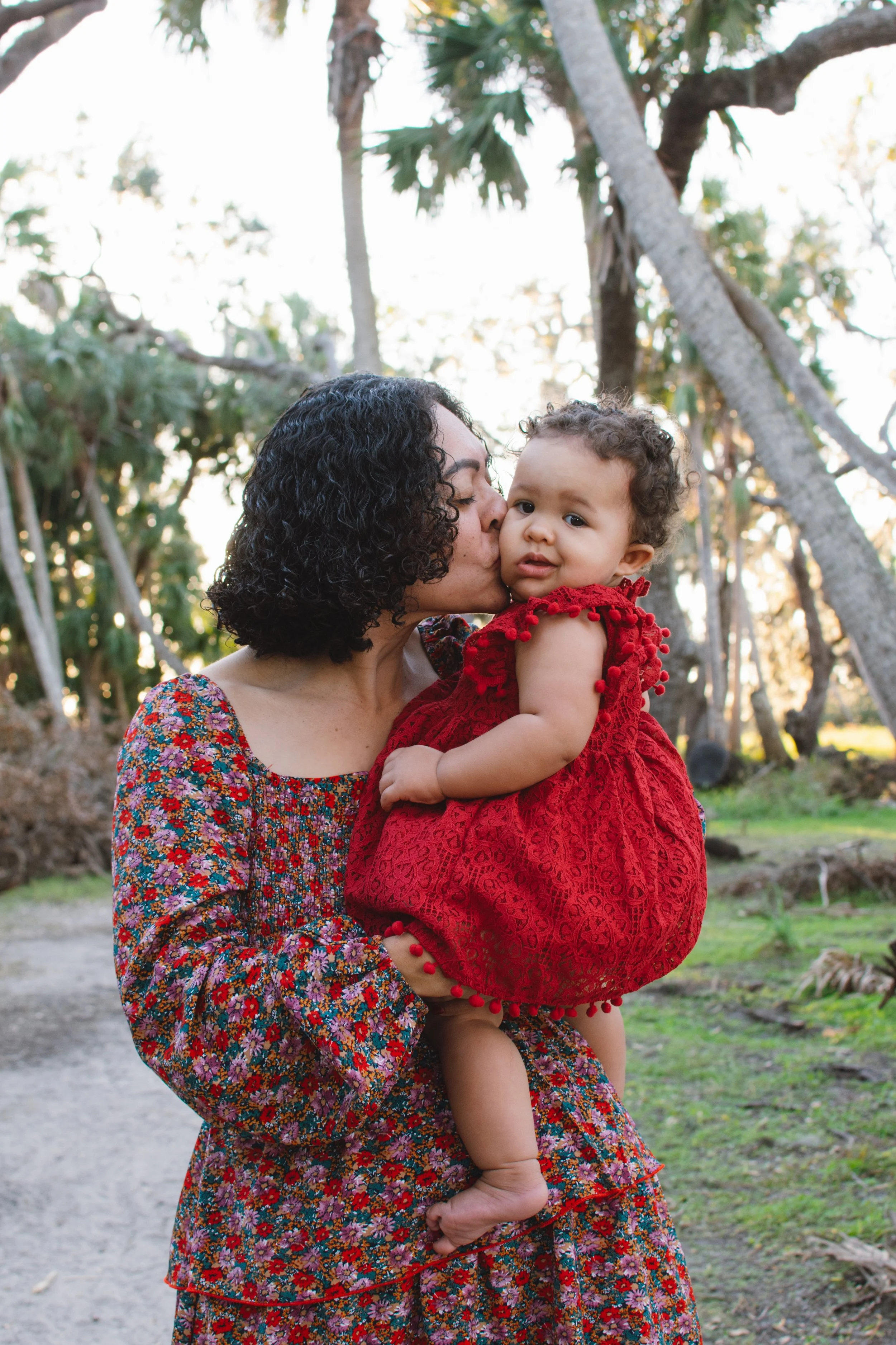 A woman with curly black hair kissing a young girl on the cheek. The girl, dressed in a red dress, has short curly hair. They are outdoors in a lush, green area with tall trees and sunlight filtering through.