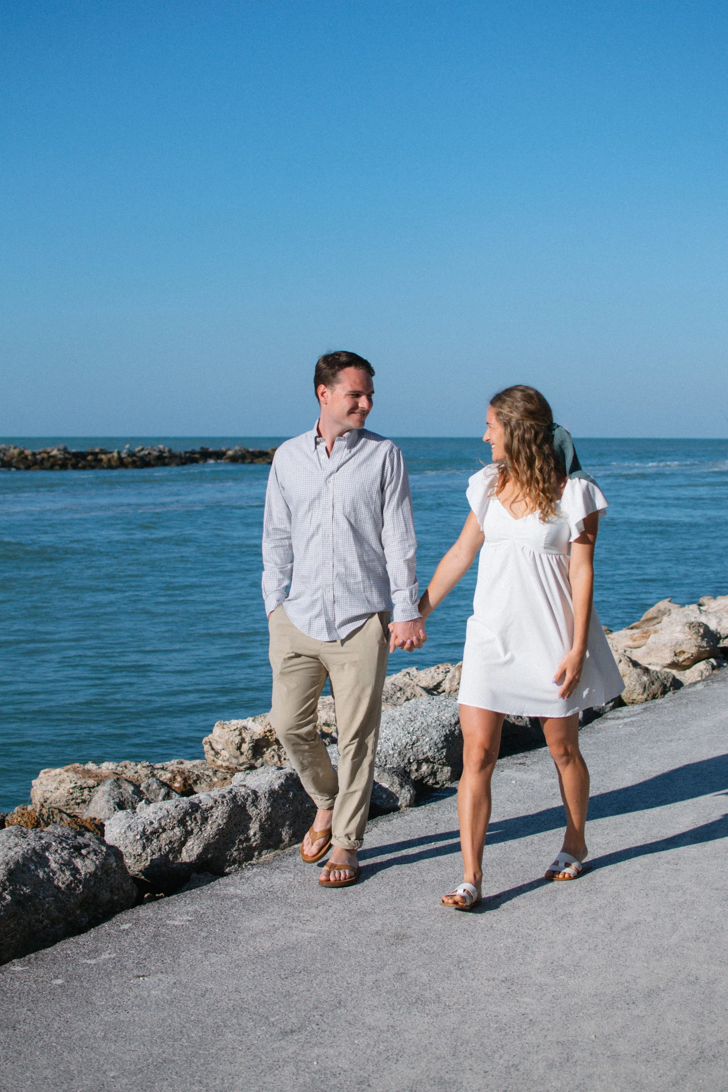 A couple holding hands and walking along a rocky path by the ocean, smiling at each other on a sunny day.
