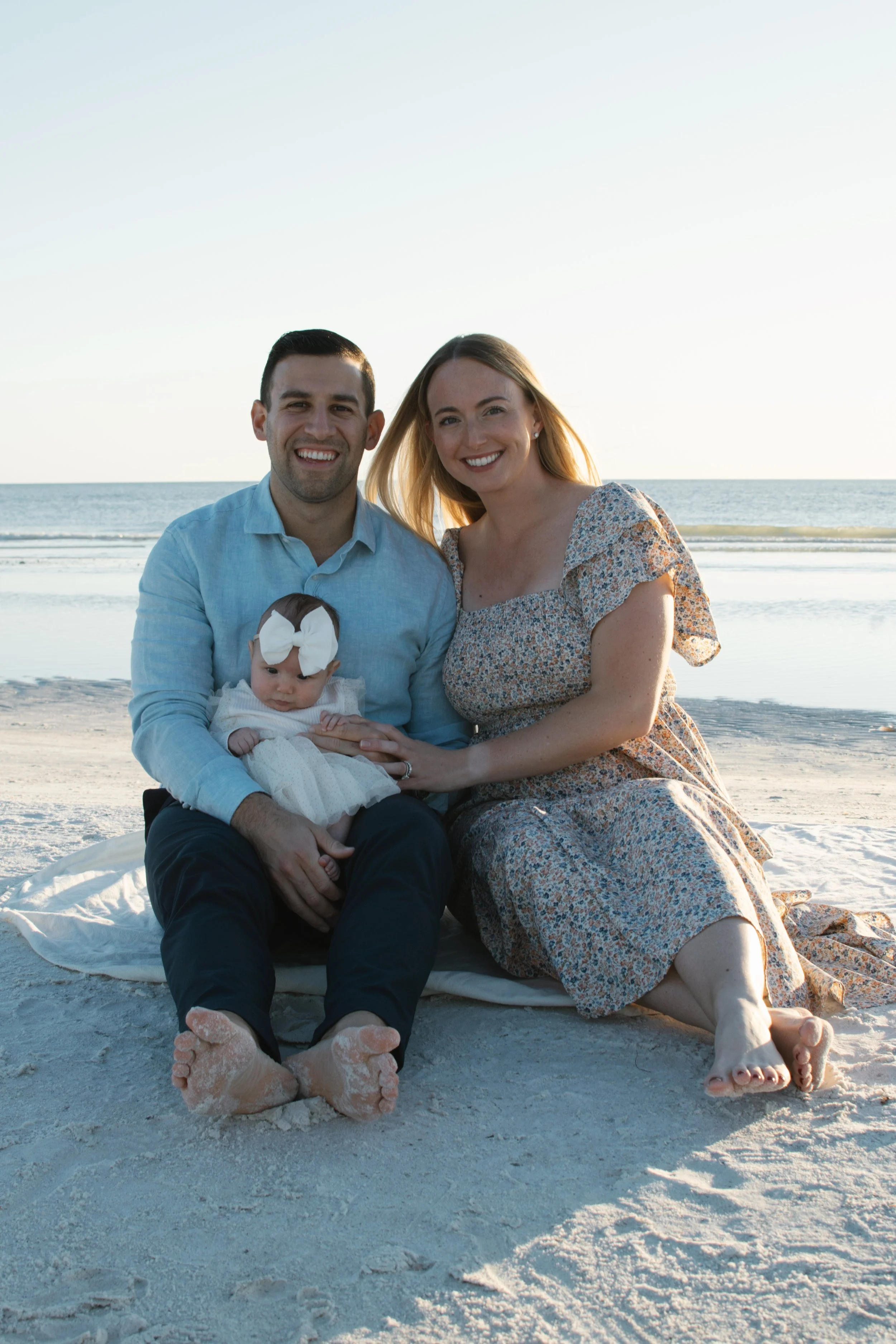 A happy family of three sitting on the beach with the ocean in the background. The father and mother are smiling and holding their baby girl who has a white bow in her hair.