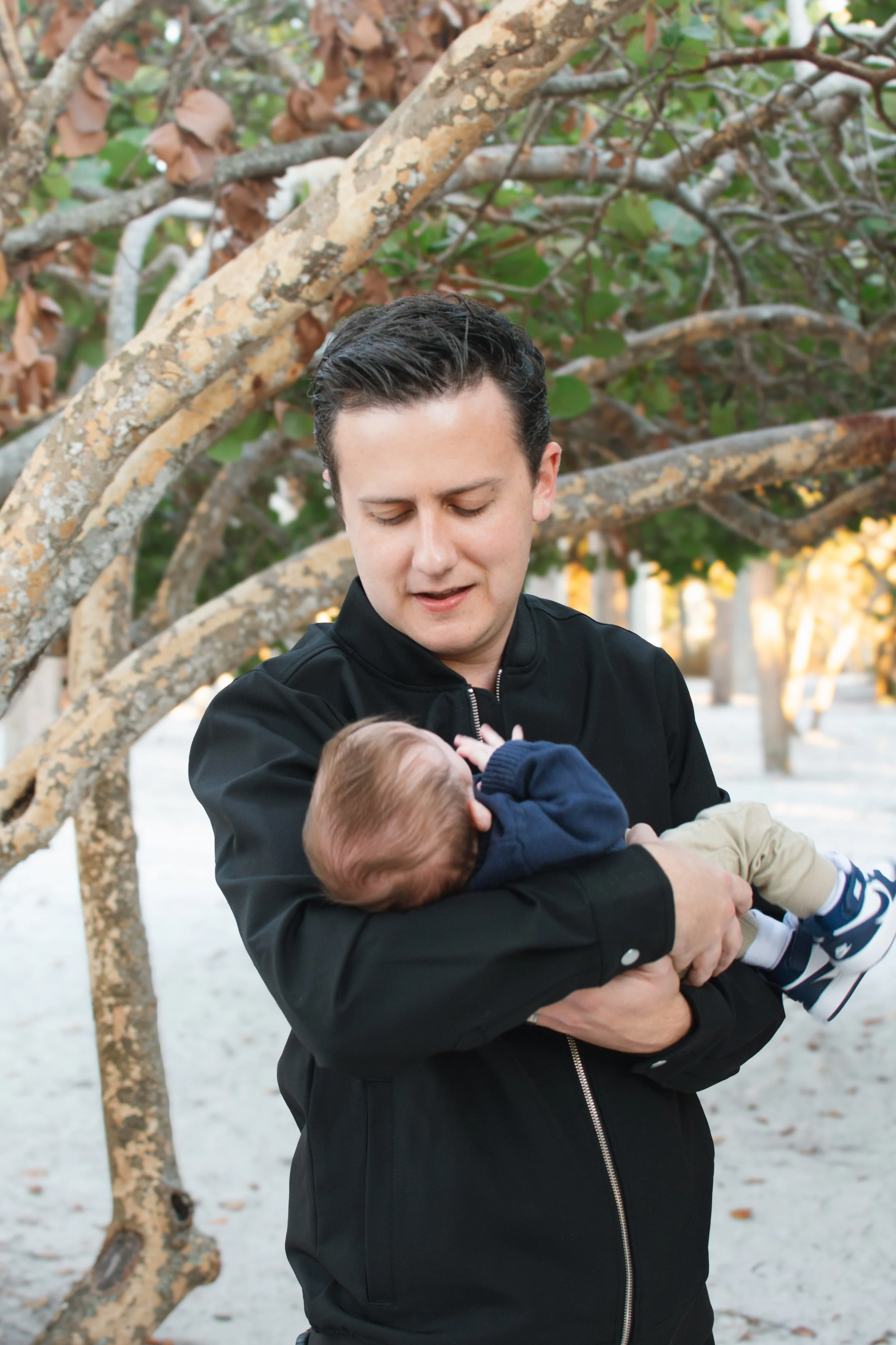 A man holding a baby in an outdoor park with trees and branches in the background.