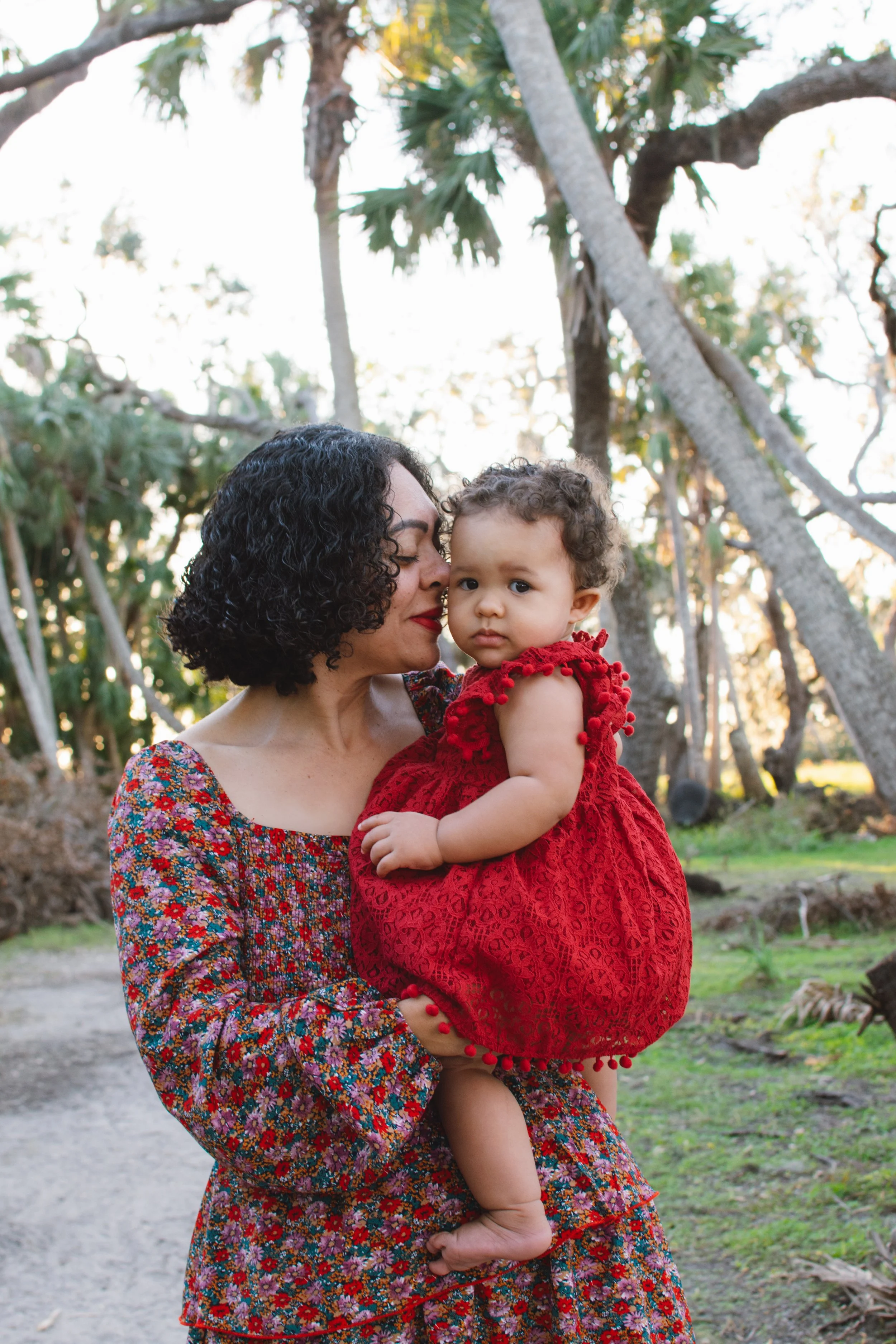 A woman holding a young girl outdoors in a wooded area with trees and sunlight in the background. The woman has curly dark hair and is wearing a colorful, patterned dress. The girl wears a red dress with pom-pom details and has curly hair, and she lo