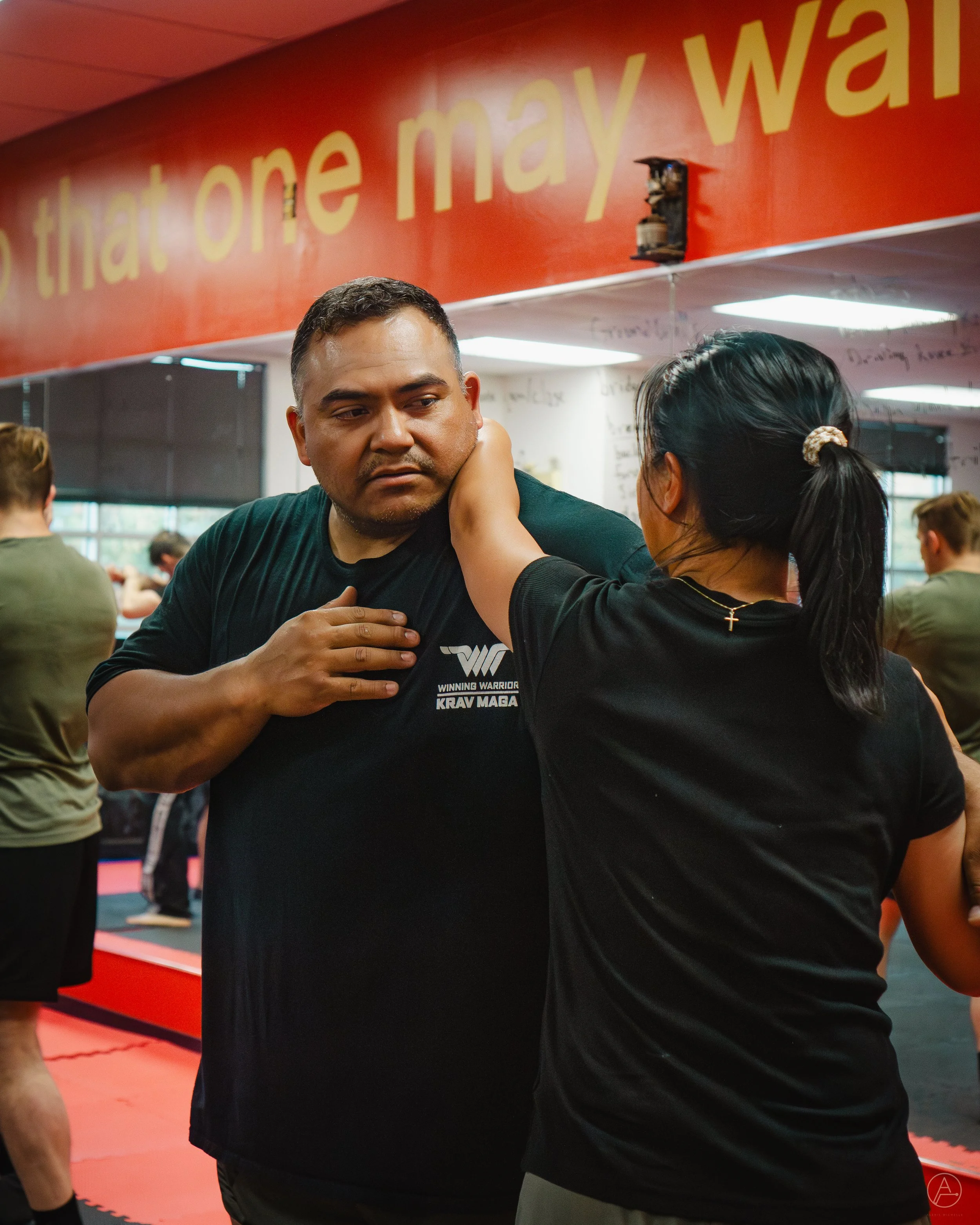 A man and woman practicing Krav Maga in a gym, with a red wall and yellow text in the background.