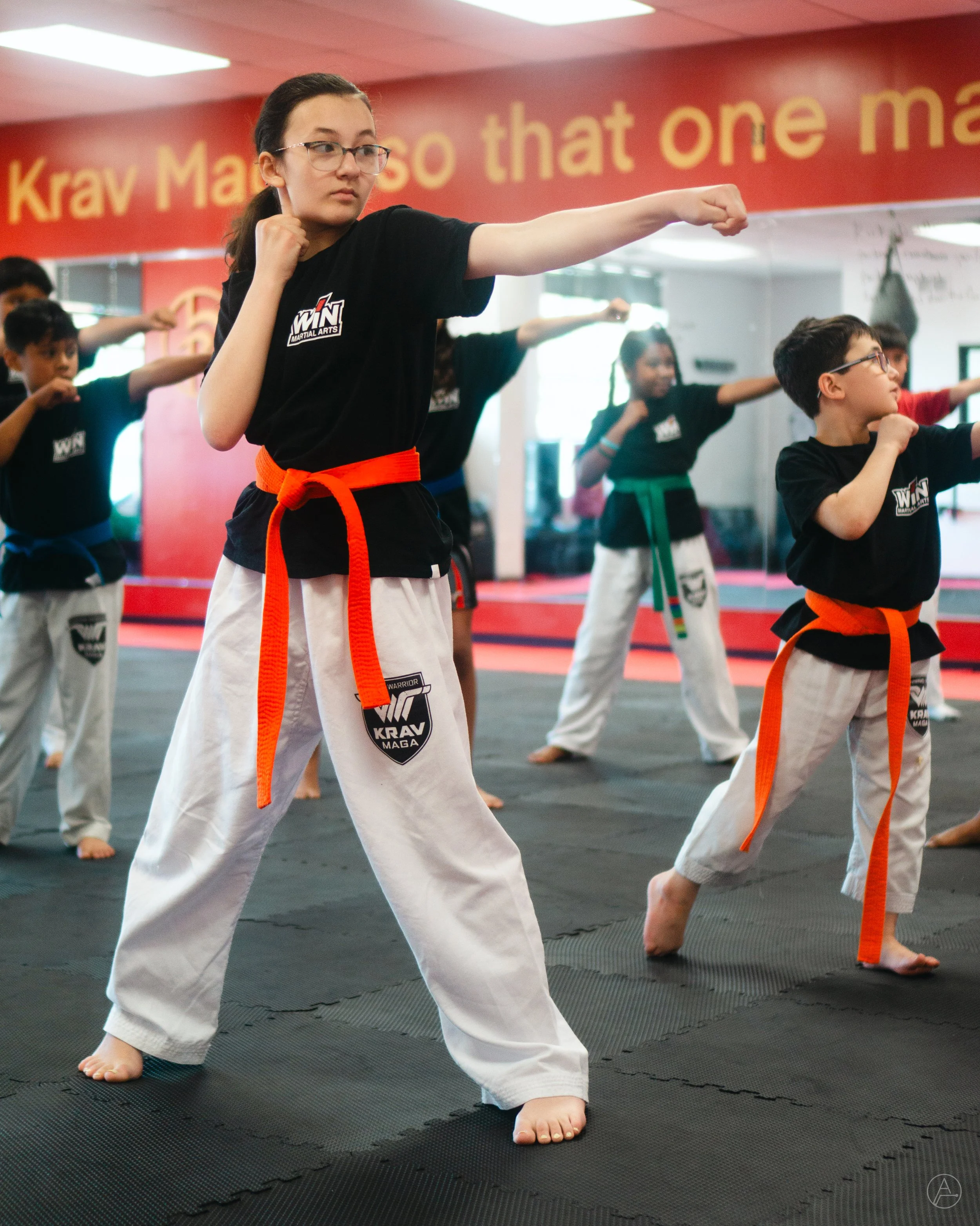 A group of children practicing martial arts in a dojo. The main girl in the foreground is throwing a punch. All are wearing martial arts uniforms with colored belts.