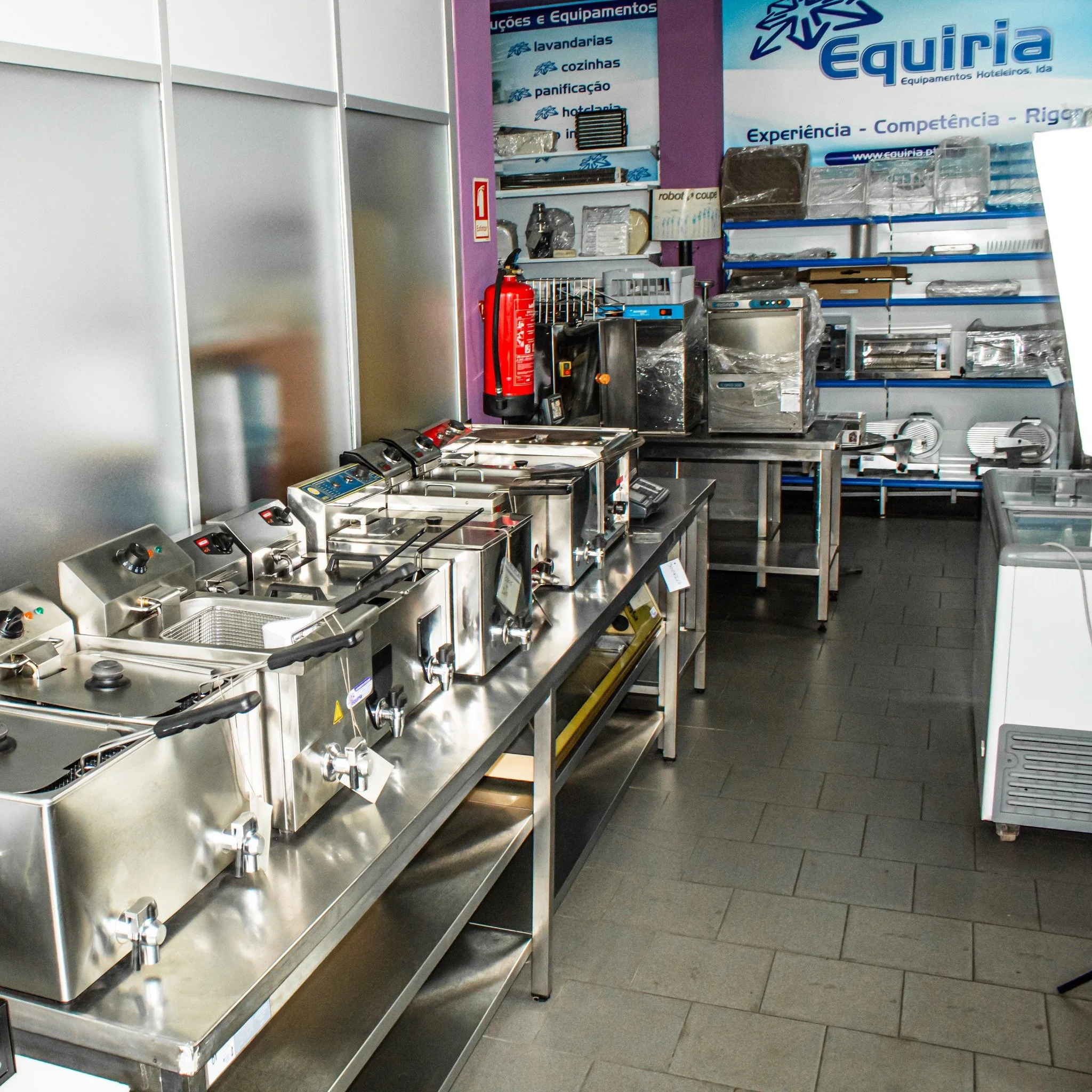 Mulher de cabelo castanho e cabelo reto, usando um chapéu bege, camiseta preta, colete preto e Jeans azul, posando ao lado de uma parede colorida amarela e vermelha com janela e mapa na parede ao fundo.
