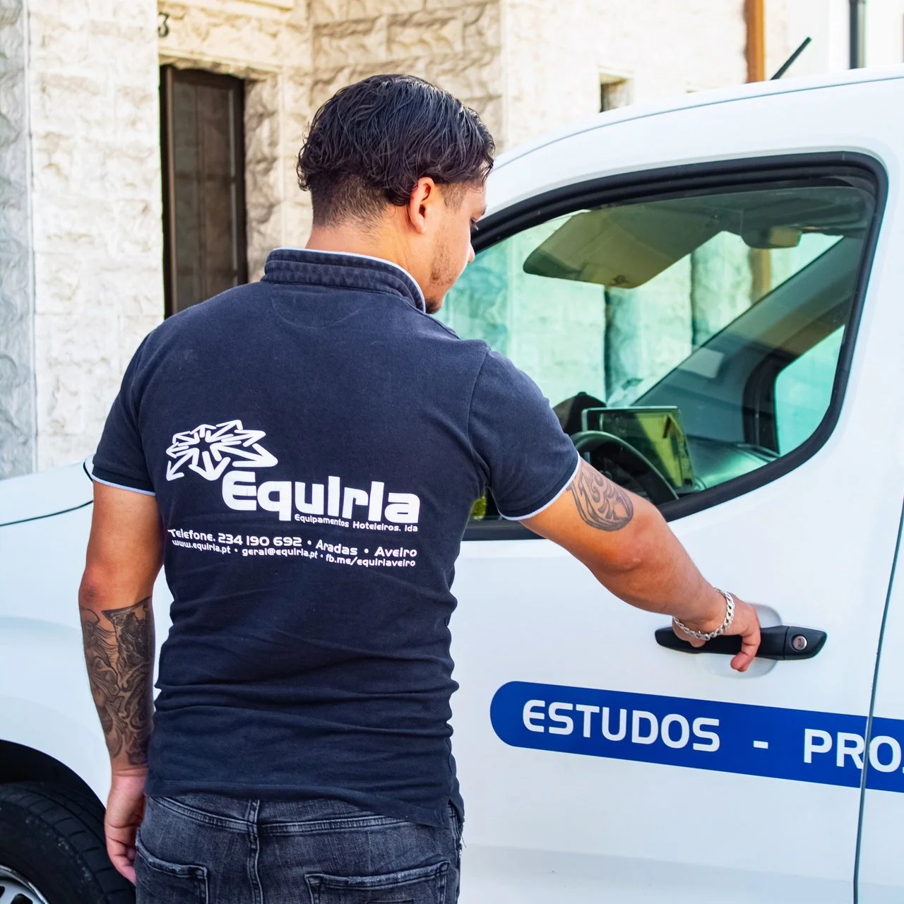 Mulher com chapéu e mochila amarela apontando para um barco tradicional de Timor-Leste, enfeitado com pinturas coloridas, ao lado de um rio em uma cidade com edifícios históricos ao fundo.