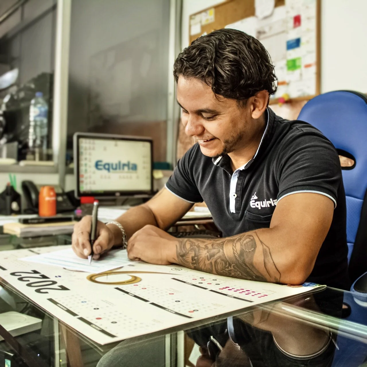 Mulher com óculos de sol transparentes, cabelo castanho escuro, brincos pequenos, colar com pingente de borboleta dourada, pele clara com sardas, usando camiseta preta.