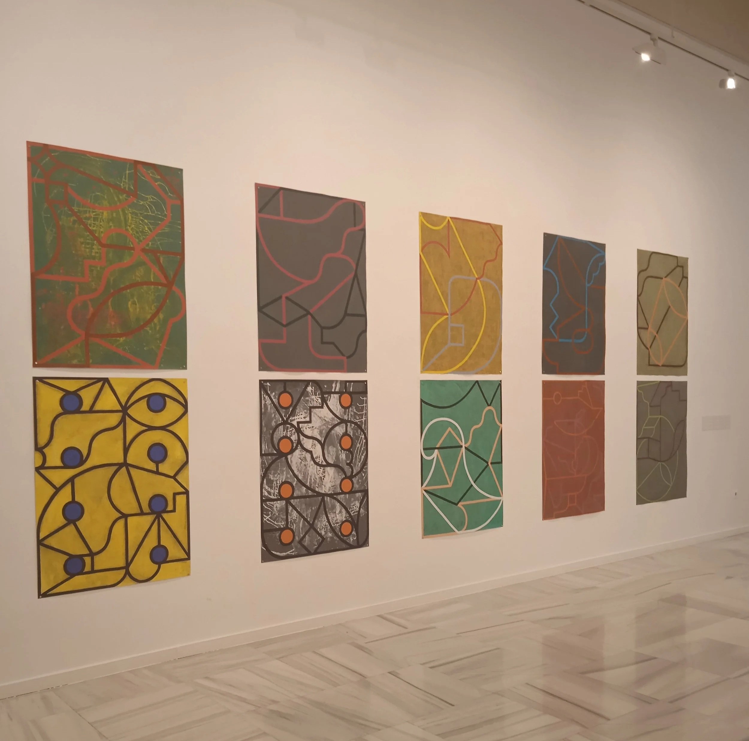 Ocho cuadros abstractos con líneas y formas de diferentes colores en una galería de arte.