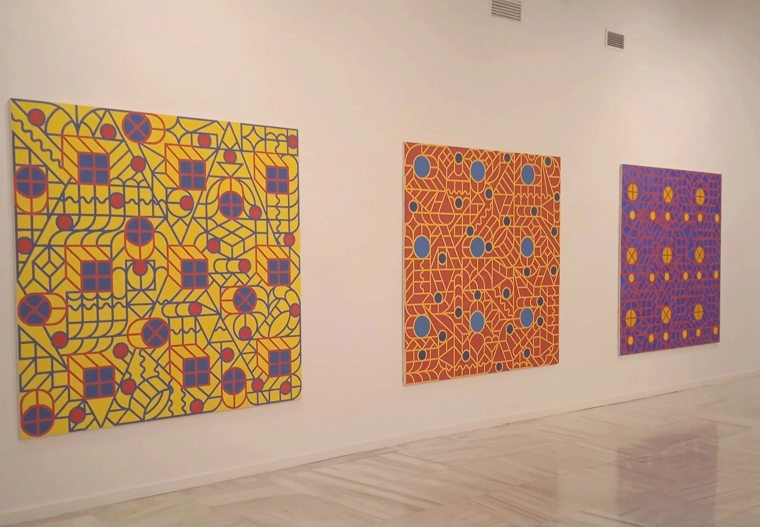 Tres cuadros con patrones geométricos y colores vibrantes en una galería de arte.