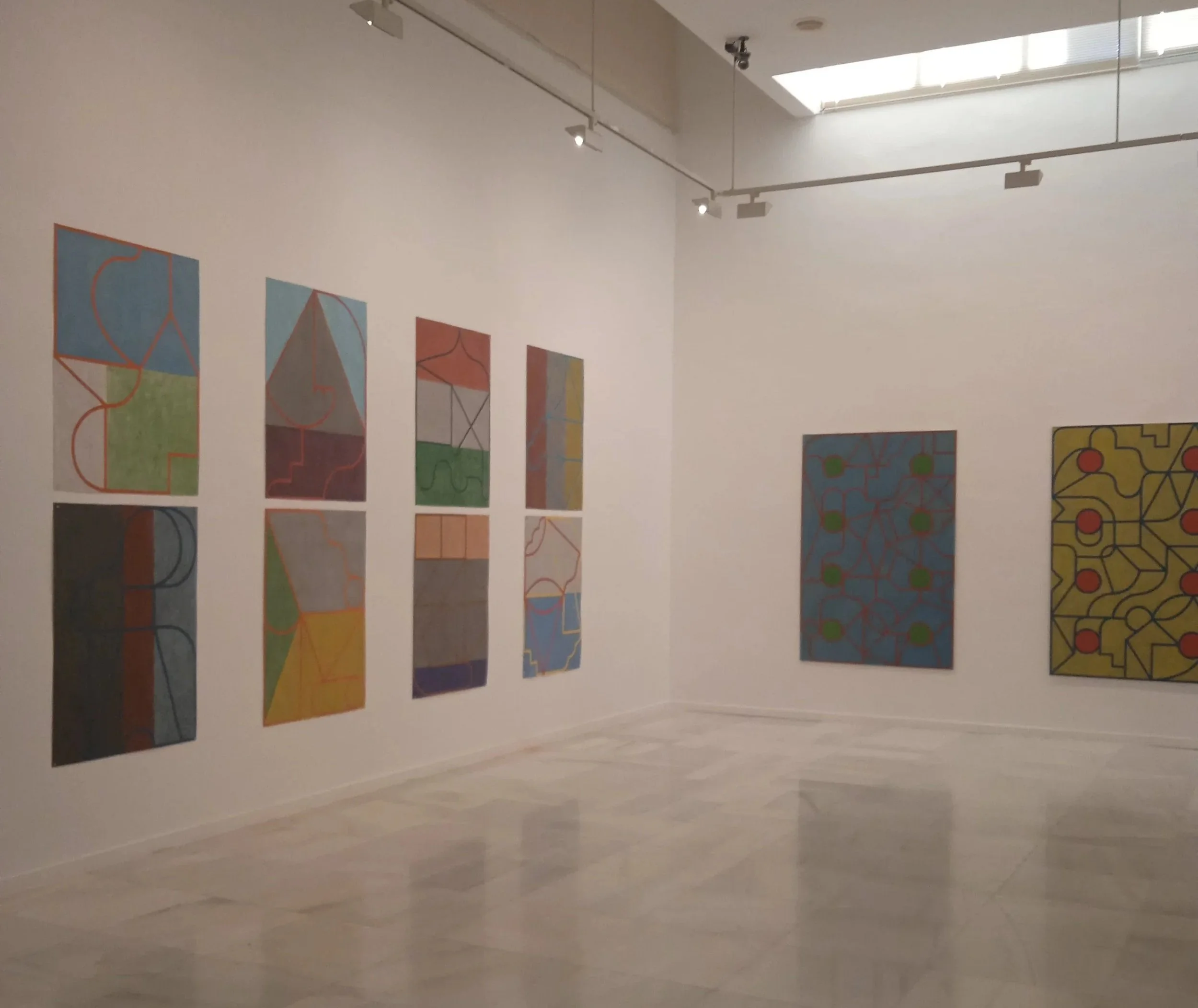 Galería de arte con nueve pinturas abstractas en la pared de fondo, enmarcadas u OT y con líneas en diferentes colores. La sala está iluminada por luz natural desde una claraboya en el techo.