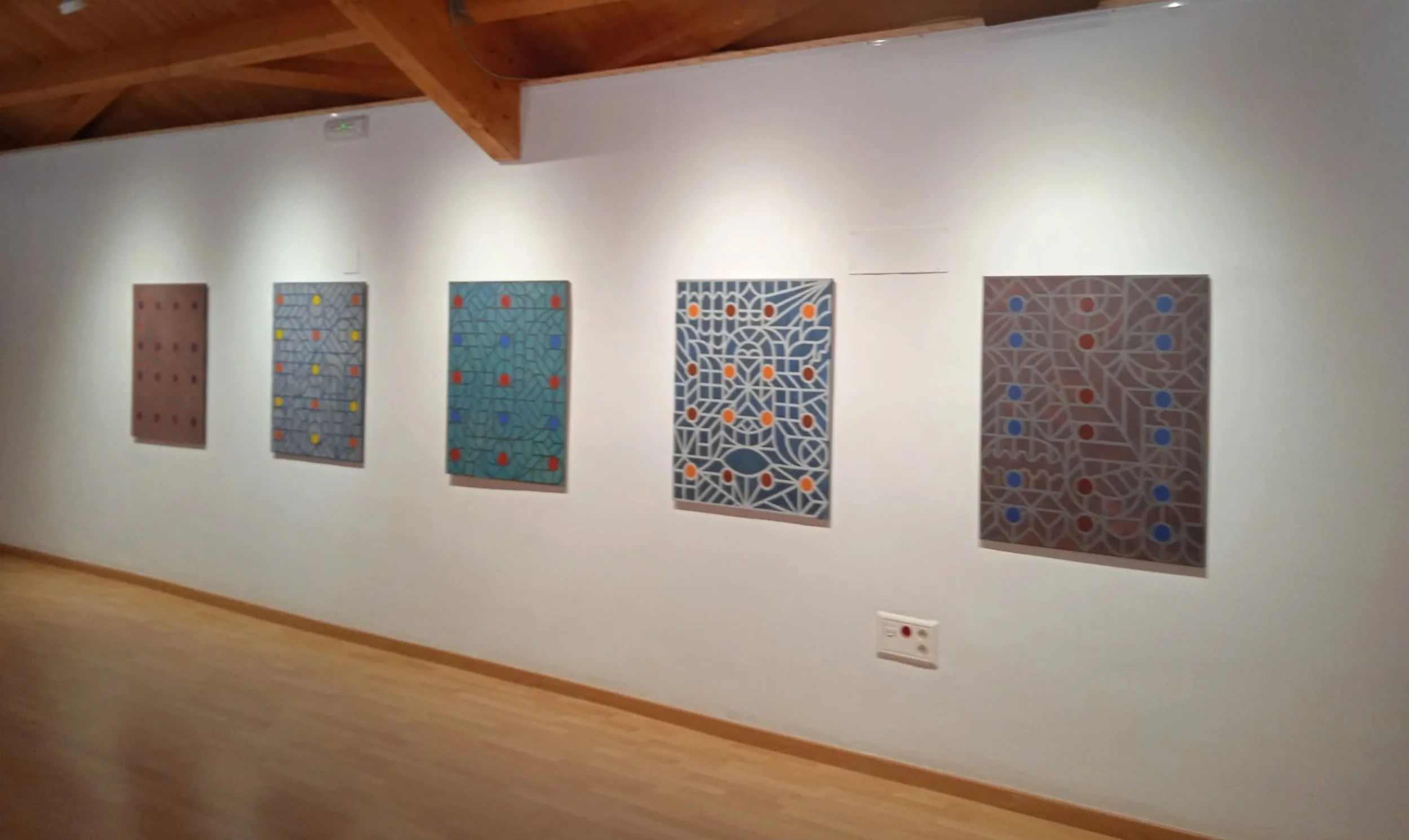 Cinco cuadros abstractos con patrones geométricos en diferentes colores colgados en una pared blanca en una galería de arte.