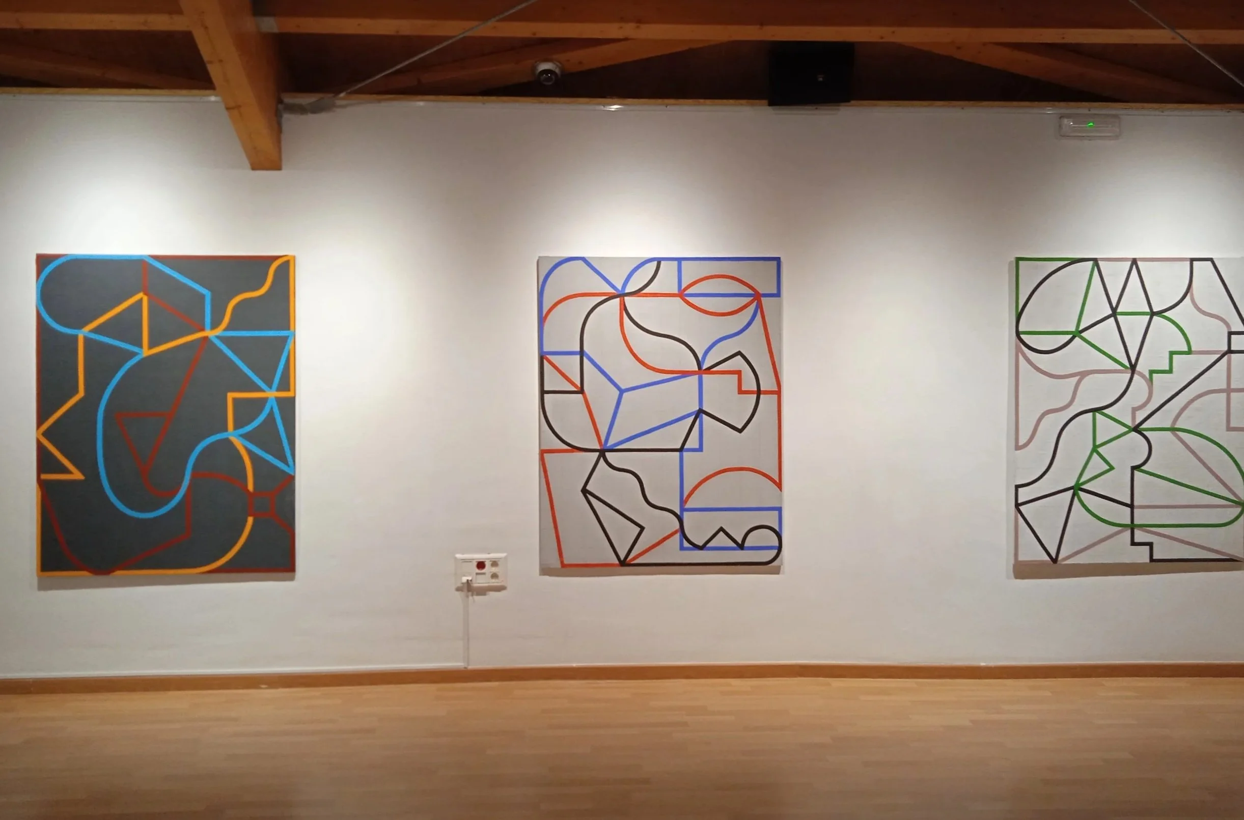 Tres pinturas de arte abstracto con líneas onduladas y formas geométricas enmarcadas de diferentes colores, colgadas en una pared de galería.