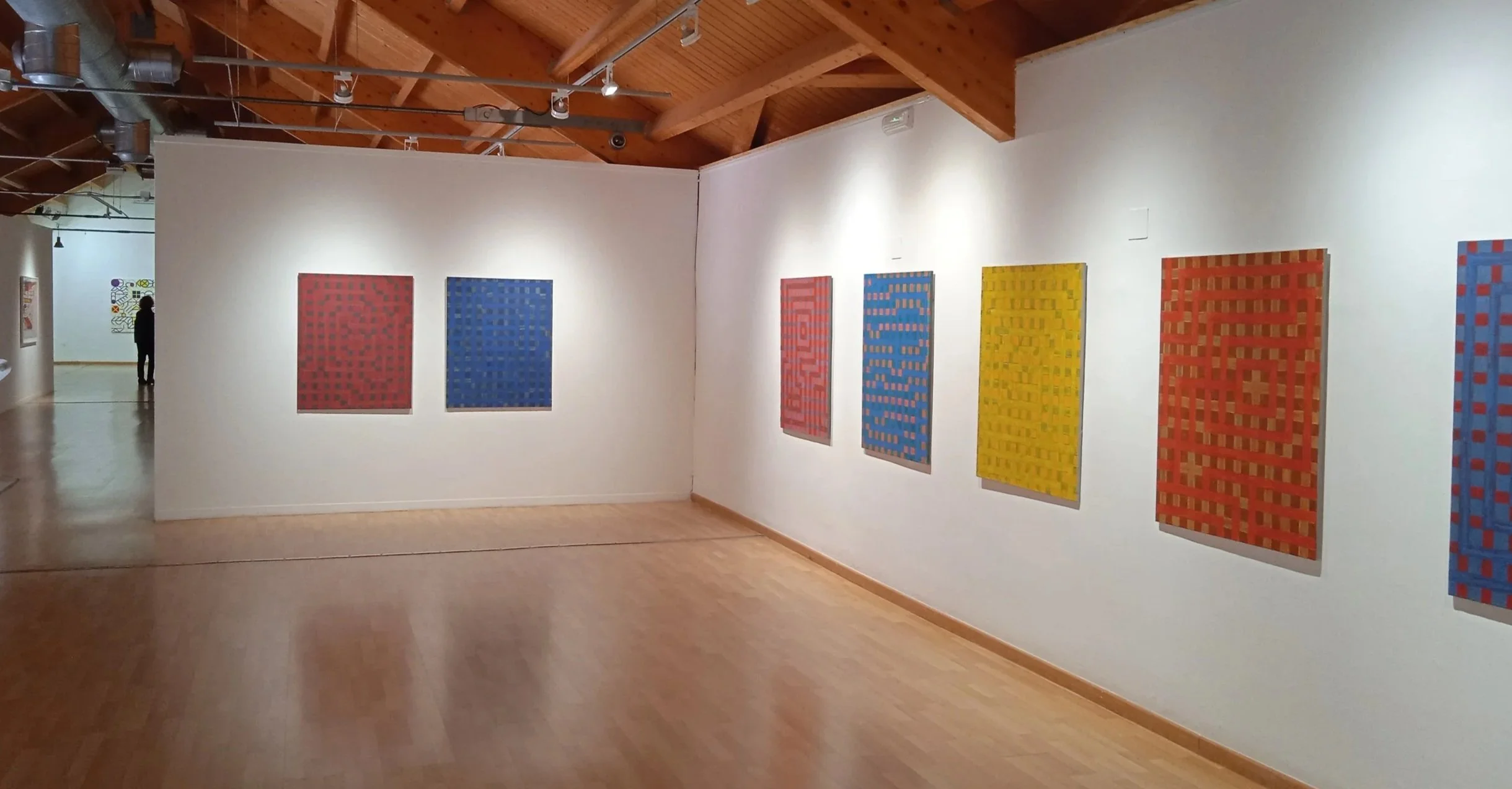 Galería de arte con cuadros abstractos de colores en una pared blanca, techo de madera y piso de madera clara.