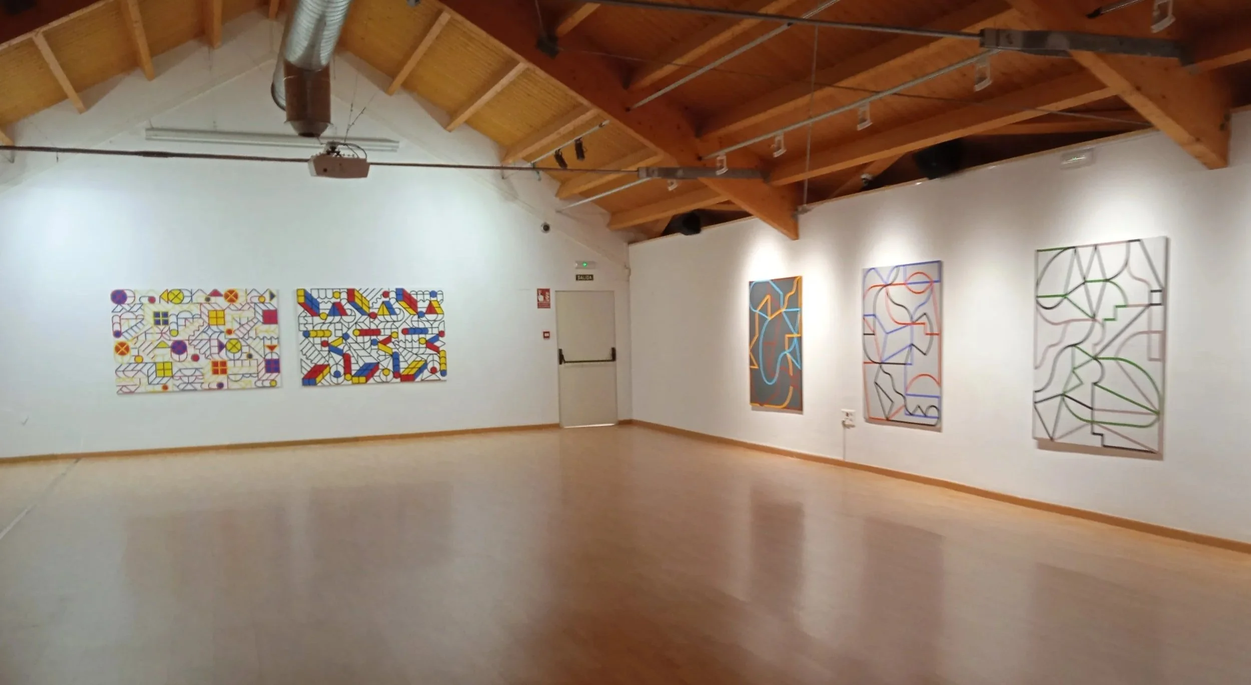 Sala de exposición con paredes blancas y techo de vigas de madera, mostrando varias obras de arte modernas y abstractas en la pared, con iluminación enfocada en cada pieza.