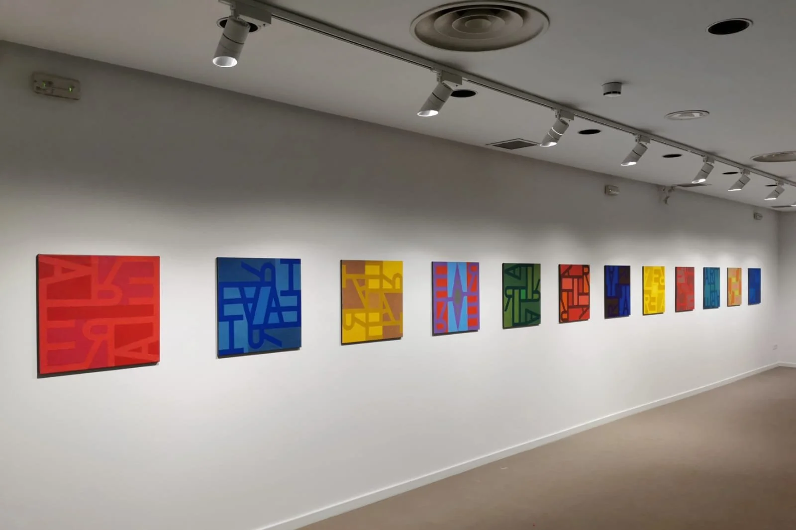 Serie de cuadros abstractos en colores variados exhibidos en una pared blanca de galería.