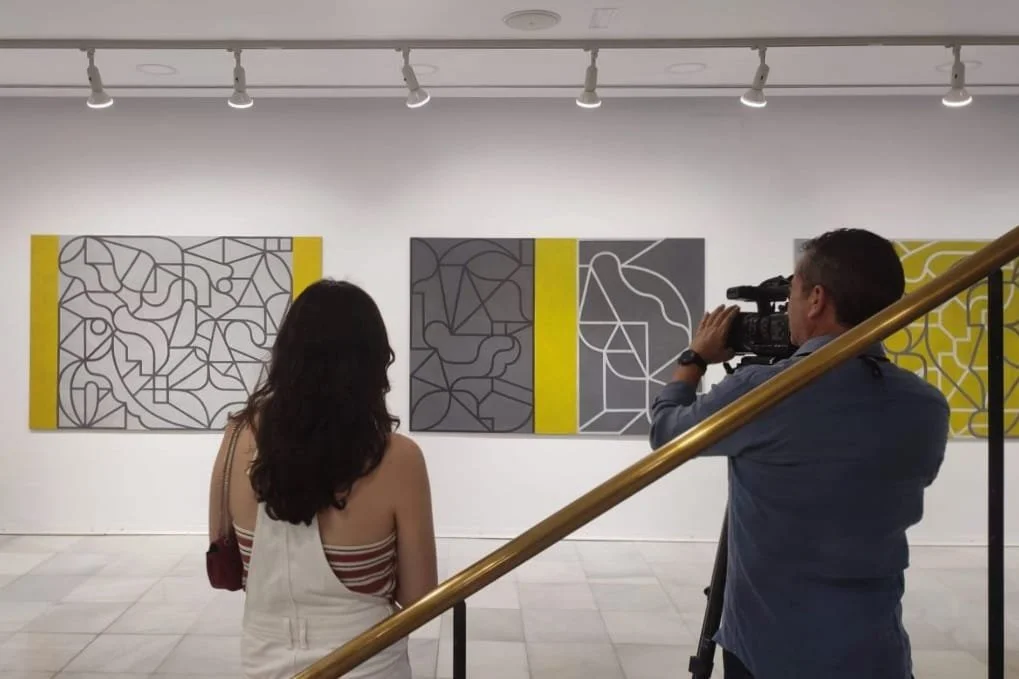 Una mujer y un hombre en una galería de arte observando y grabando pinturas abstractas con líneas negras y fondos grises y amarillos.