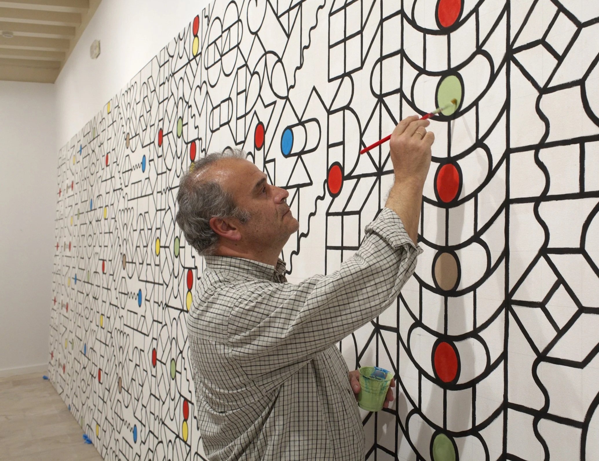 Hombre pintando un mural abstracto con formas geométricas y colores en una pared blanca en un espacio interior.