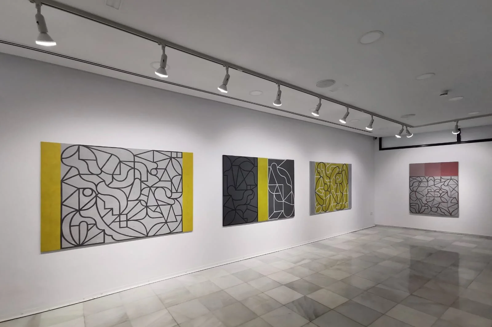 Galería de arte con cinco pinturas abstractas en lienzos colgados en una pared blanca, con iluminación dirigida desde arriba.